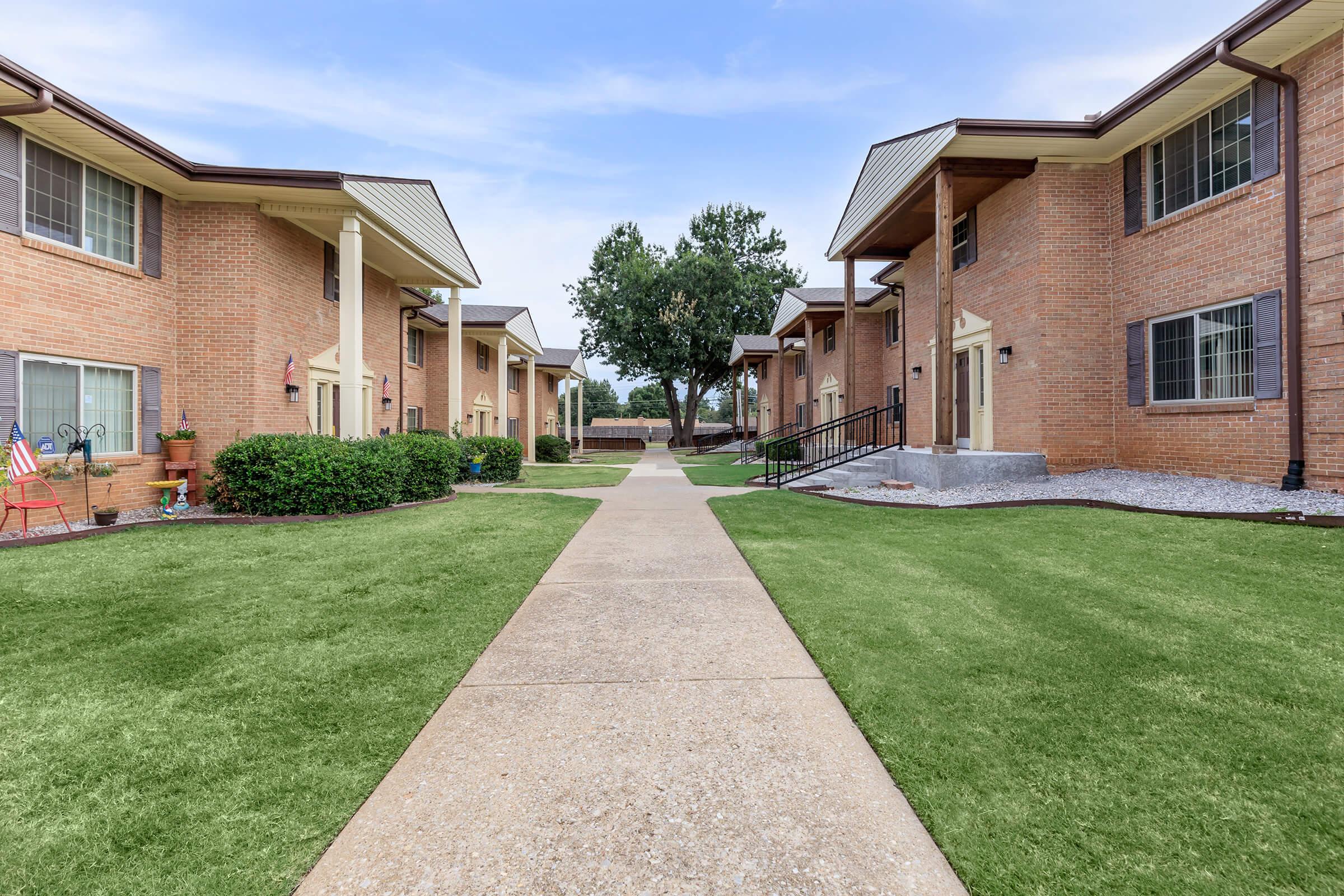 A well-maintained apartment complex featuring brick buildings along a clean pathway. Lush green grass and trees are visible, along with decorative plants and an American flag near one of the entrances. The overall setting is inviting and orderly.