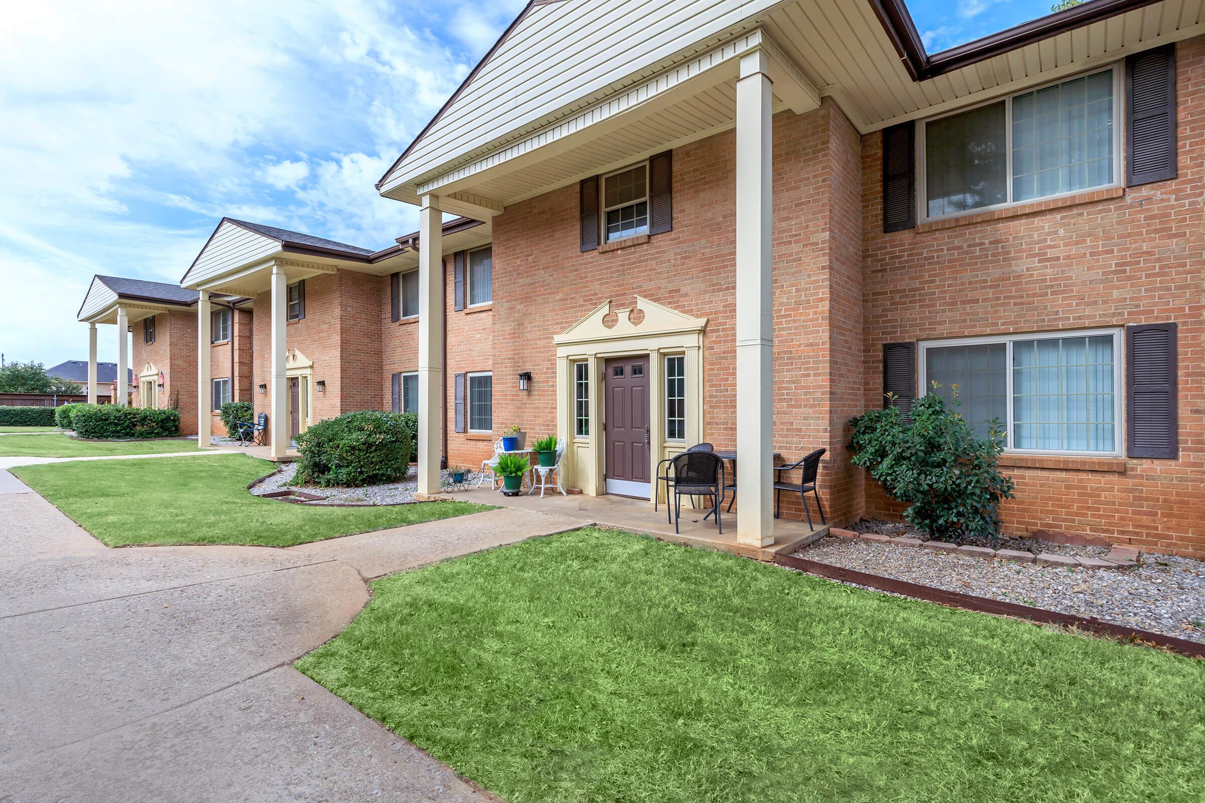 A brick apartment building with multiple units. The entrance features a decorative arch and a door with two chairs beside it. Well-maintained grass and landscaping surround the pathways leading to the doors, under a partly cloudy sky.