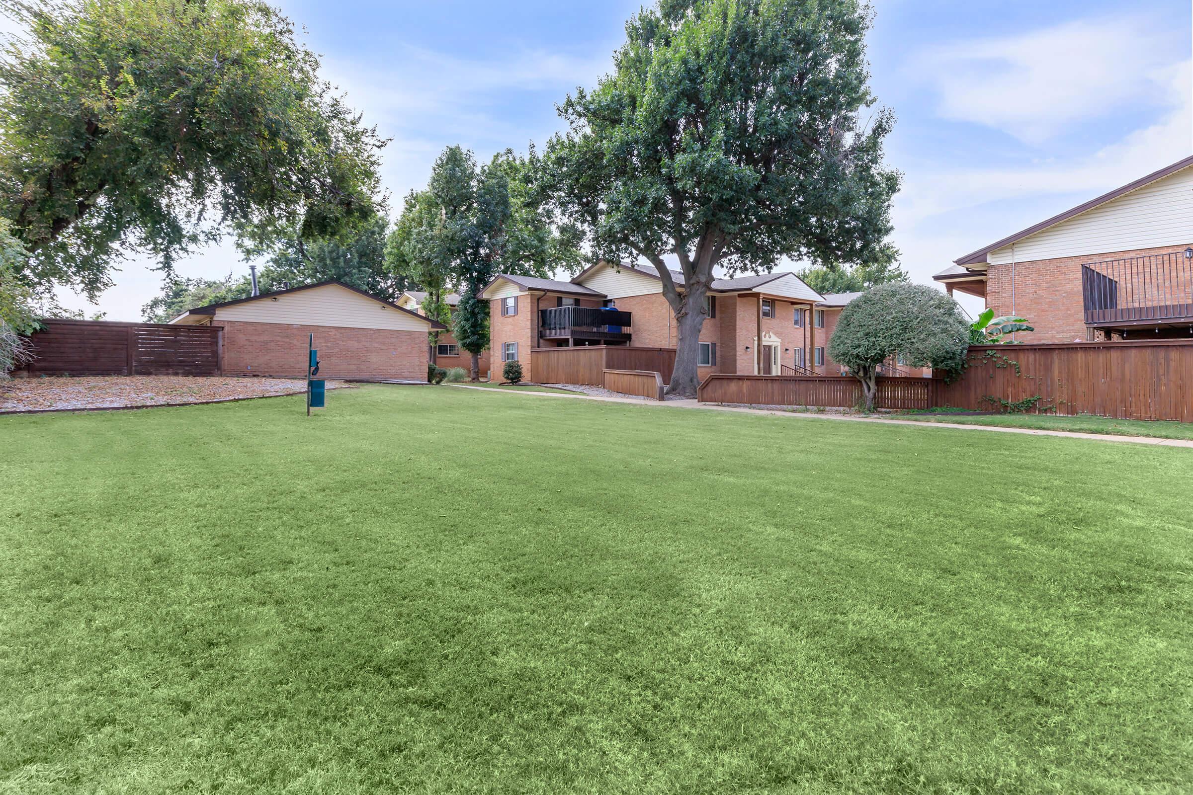 Open backyard area with a well-maintained green lawn, surrounded by residential houses. Trees provide shade, and a small wooden fence separates the properties. The sky is bright and clear, creating a pleasant outdoor atmosphere.