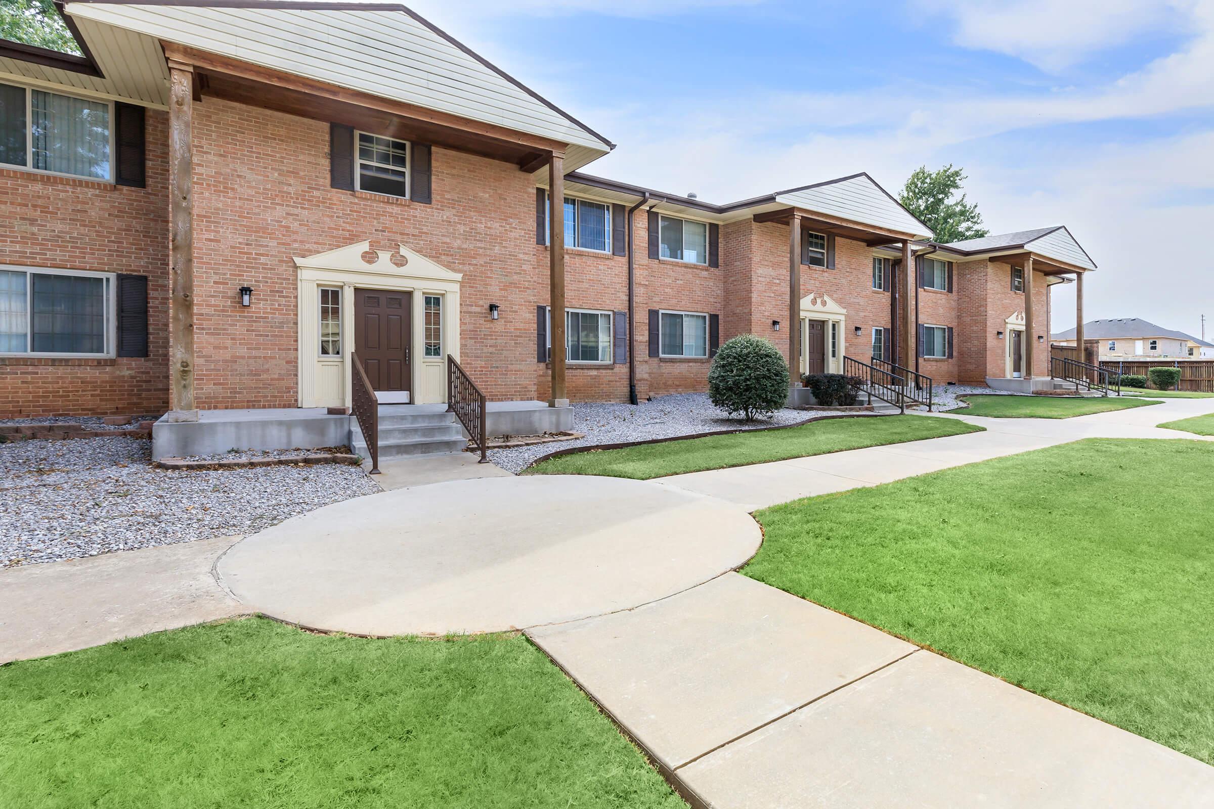 Brick apartment buildings with two stories, featuring wood accents and landscaped lawns. A paved walkway curves around the landscaped area, leading to the entrance doors of the units. The sky is clear with some clouds, and there are small bushes near the buildings.