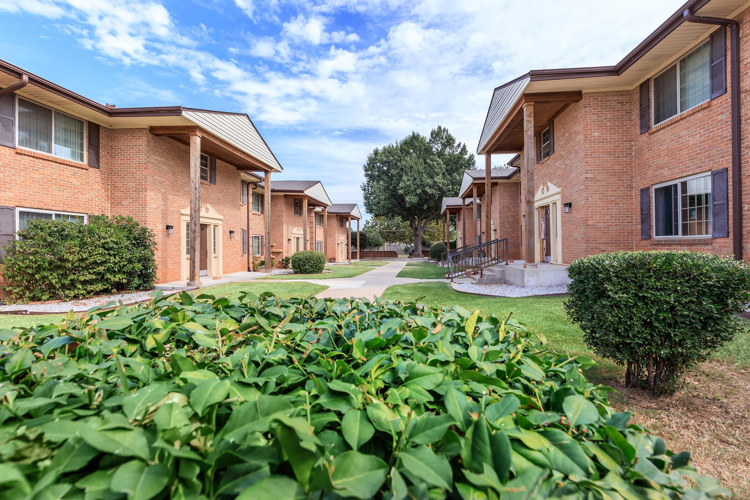 A view of a residential complex with brick buildings and green landscaping. Pathways connect the units, and there are bushes and trees in the foreground. The sky is partly cloudy, creating a pleasant outdoor atmosphere.