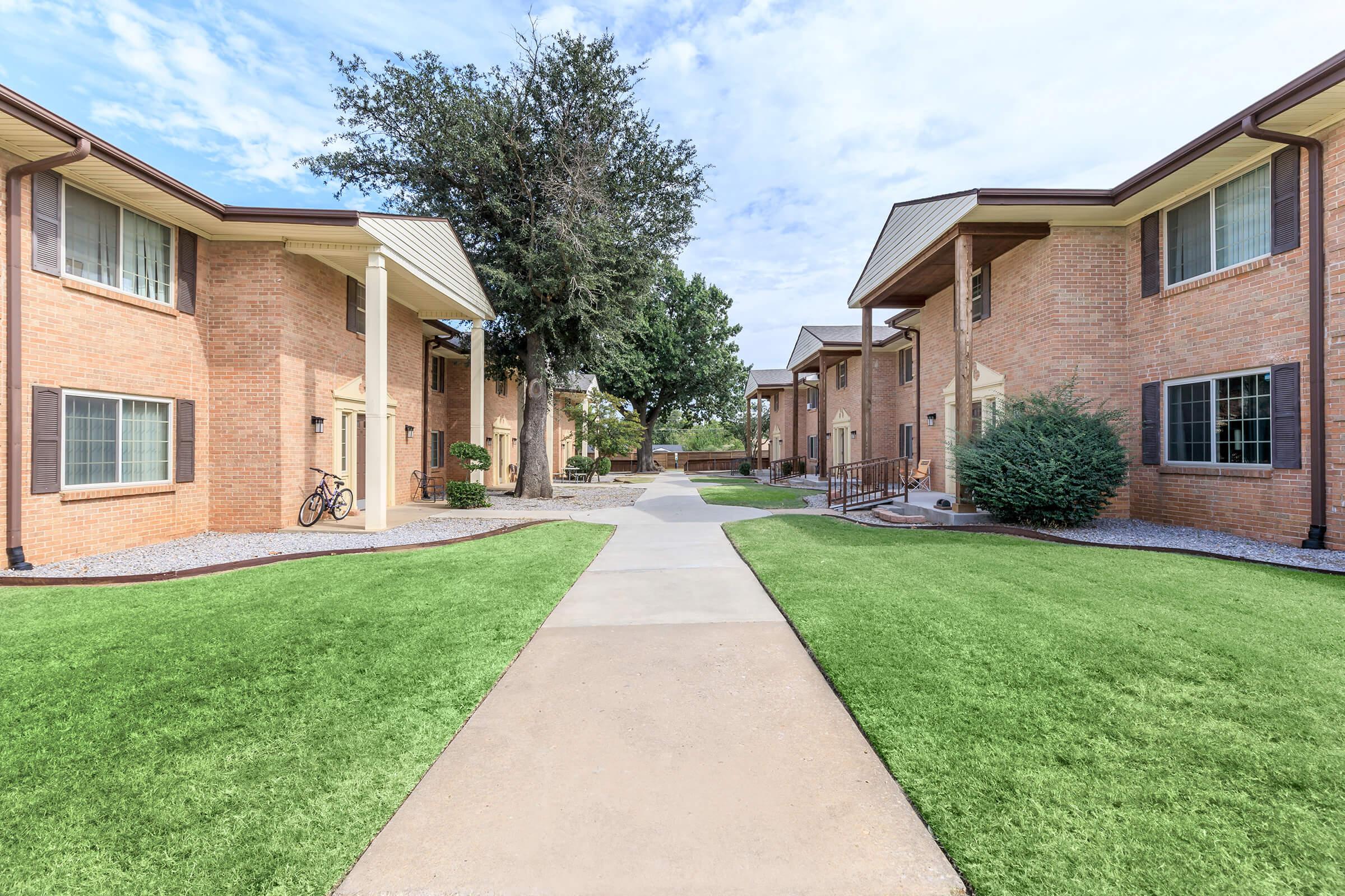 A landscaped pathway runs between two brick apartment buildings. Lush green grass lines the walkways, with a bike parked near one building. Trees and shrubs provide a natural backdrop under a blue sky with scattered clouds. The scene conveys a peaceful residential environment.