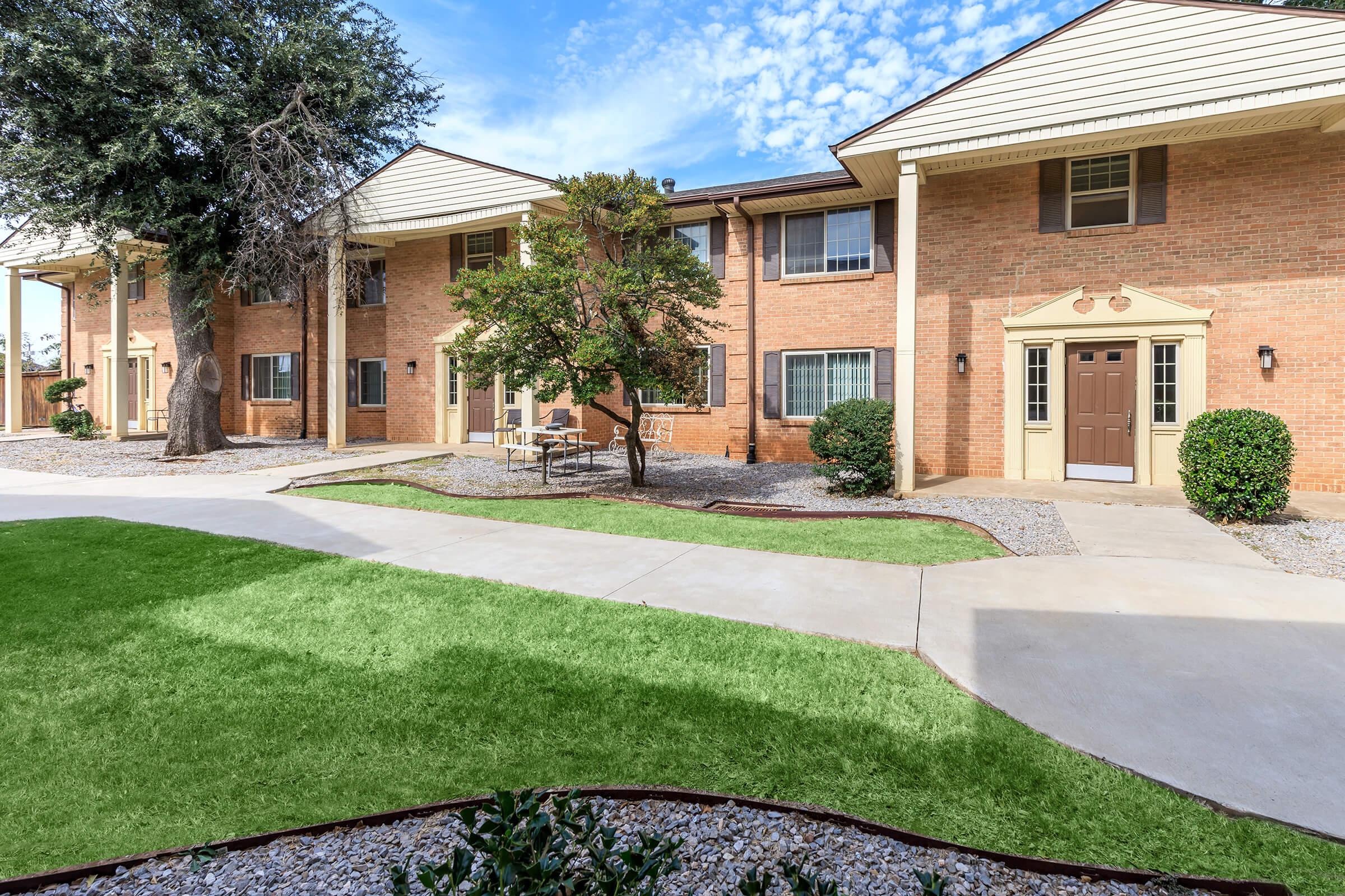 A well-maintained multi-unit brick apartment building surrounded by green grass and landscaping. The pathway leads to the entrance of the building, with seating areas visible in the courtyard. The sky is partly cloudy, adding a bright and inviting atmosphere.