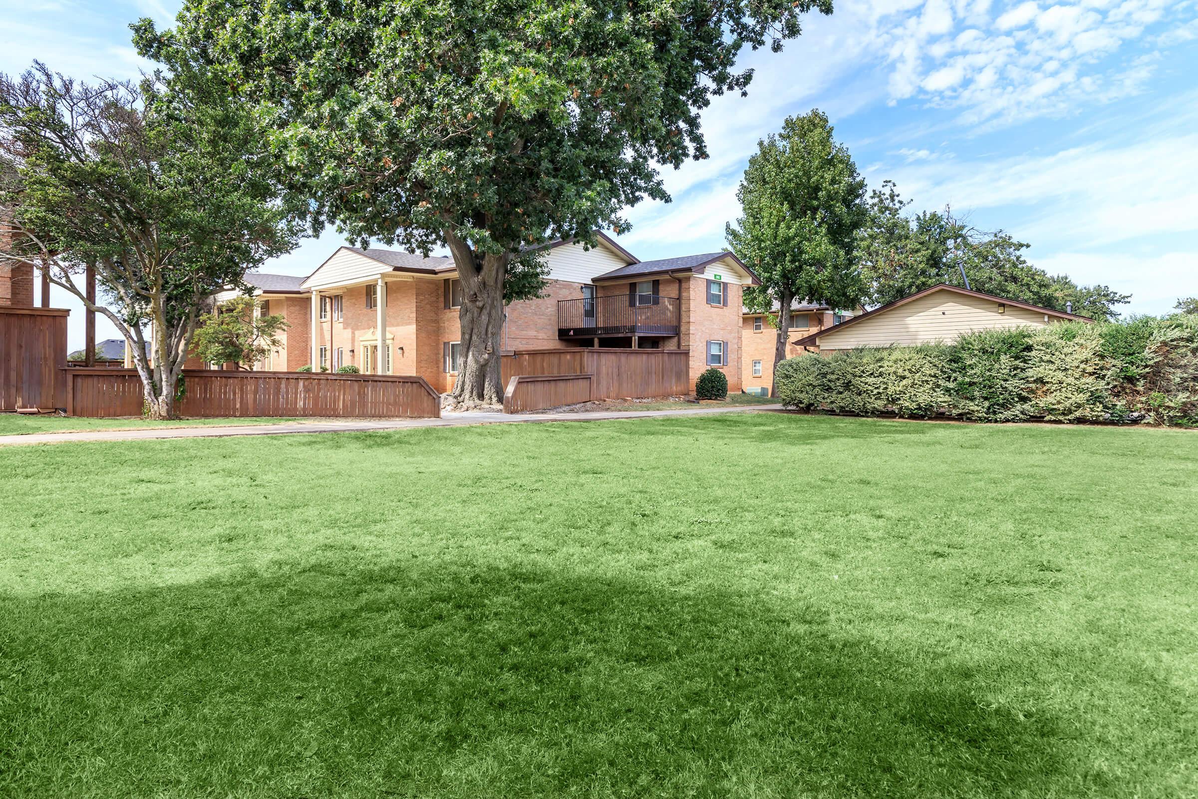 A well-maintained grassy lawn surrounded by trees and shrubs, with several residential buildings in the background. The sky is partly cloudy, creating a bright and inviting atmosphere. The scene conveys a peaceful suburban setting.