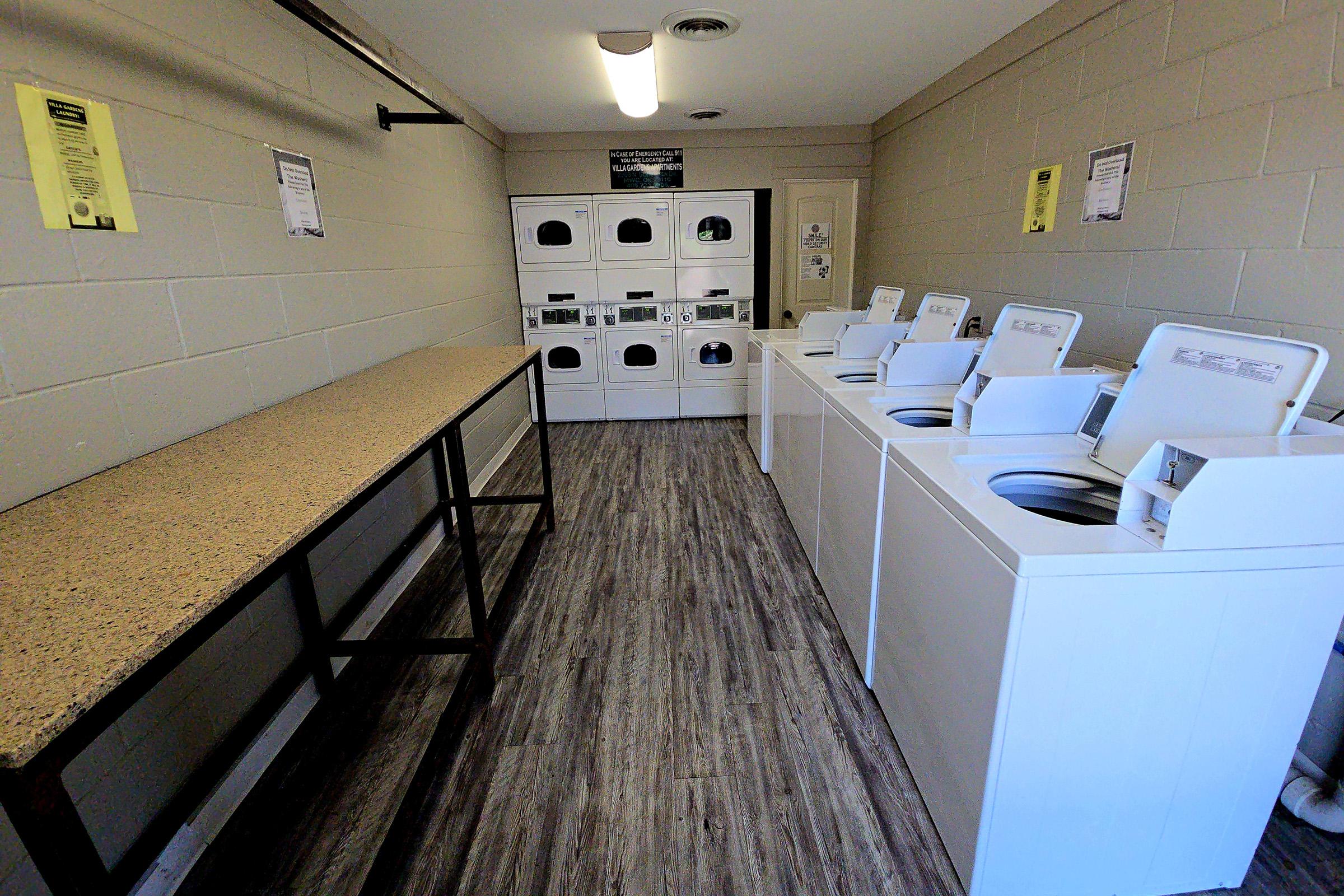 A clean and organized laundry room featuring multiple white washing machines and dryers arranged in a row. There is a long countertop made of light-colored stone for folding clothes, and notices are posted on the walls. The flooring is a wooden laminate finish.