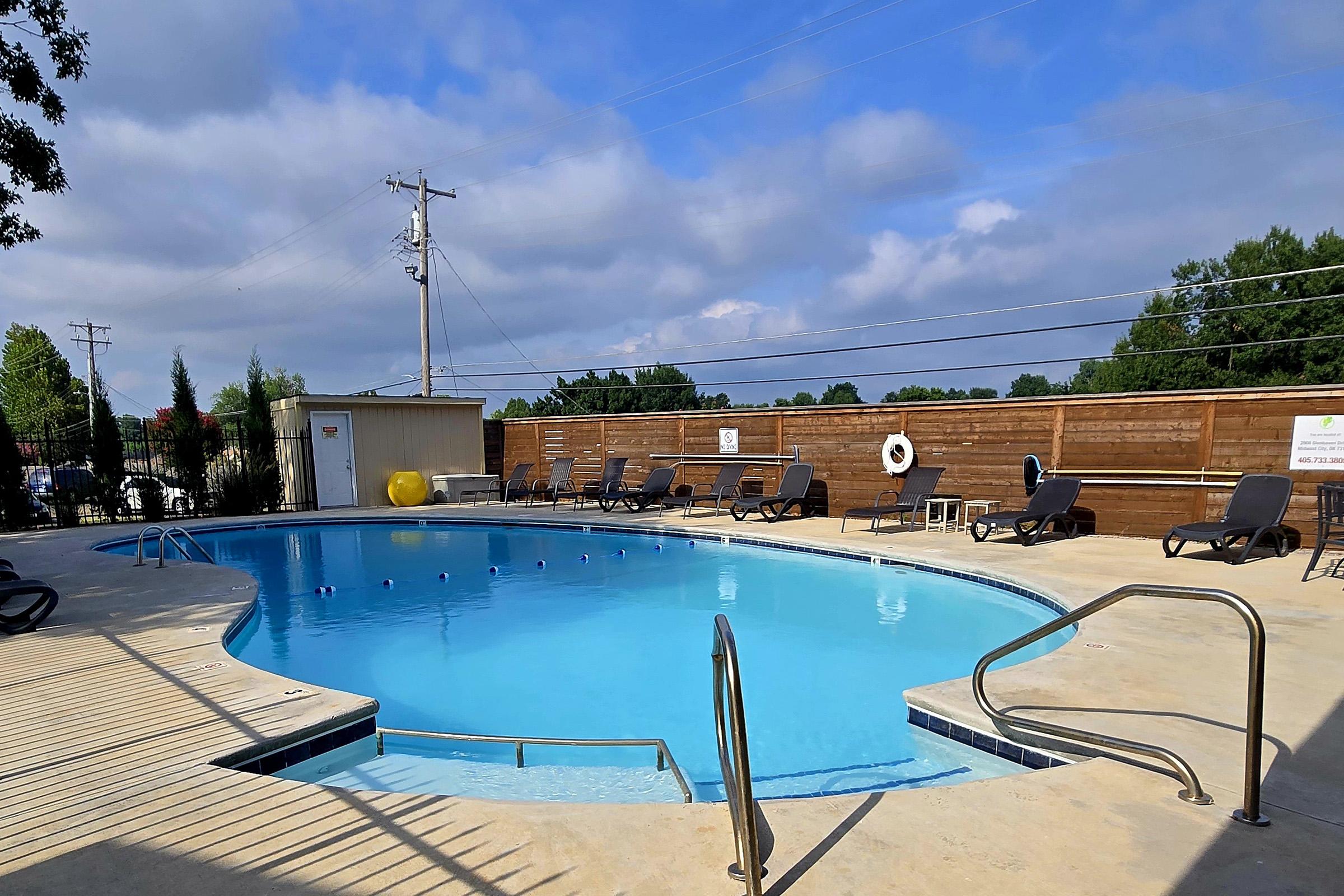 A clear blue swimming pool surrounded by lounge chairs and a wooden fence. There are utility poles in the background and a small building nearby. The sky is partly cloudy, creating a bright and inviting atmosphere.
