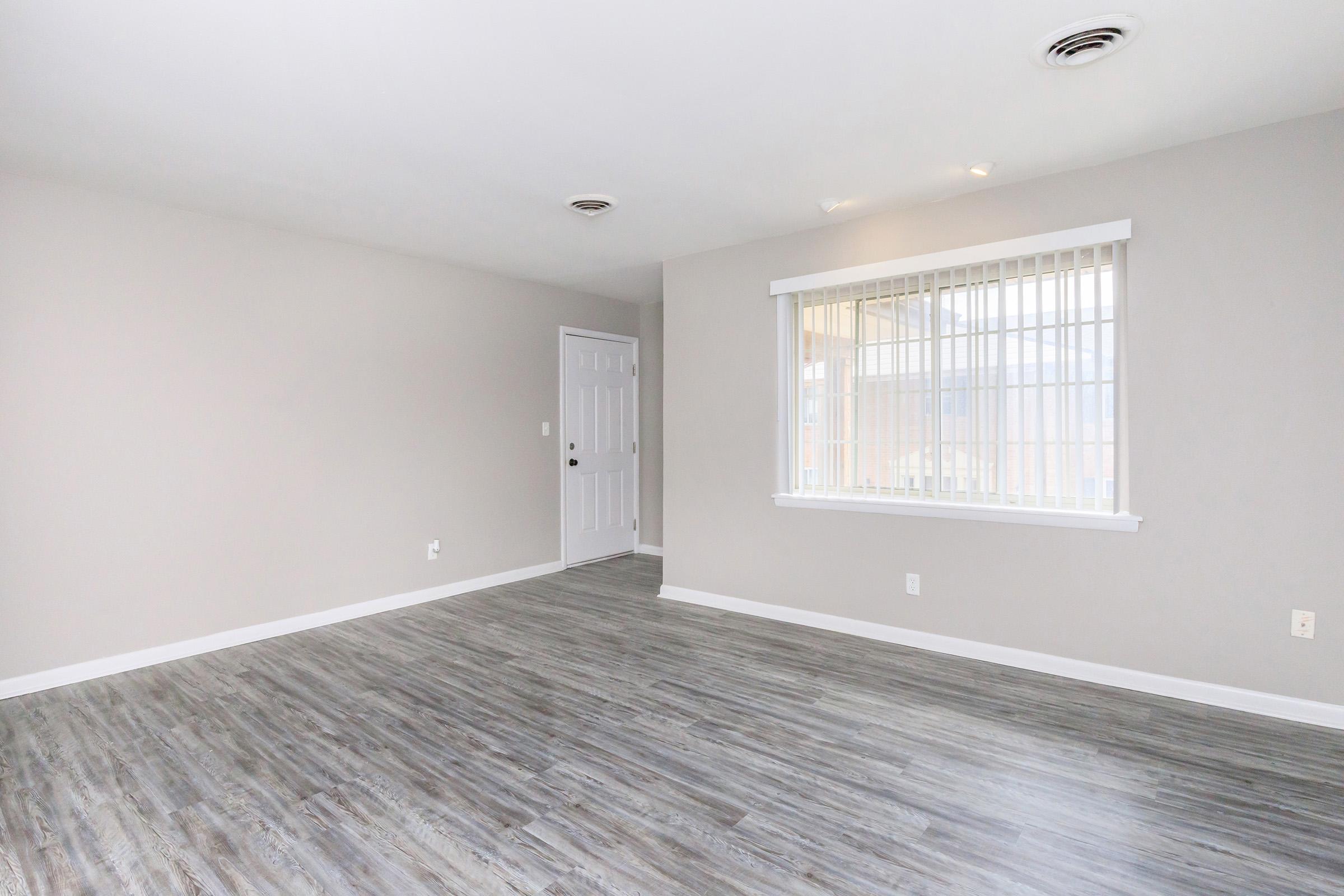 A vacant room with light gray walls and flooring, featuring a white window with blinds on the left side. A closed white door is visible to the right. The space is well-lit and empty, creating a clean and modern atmosphere.