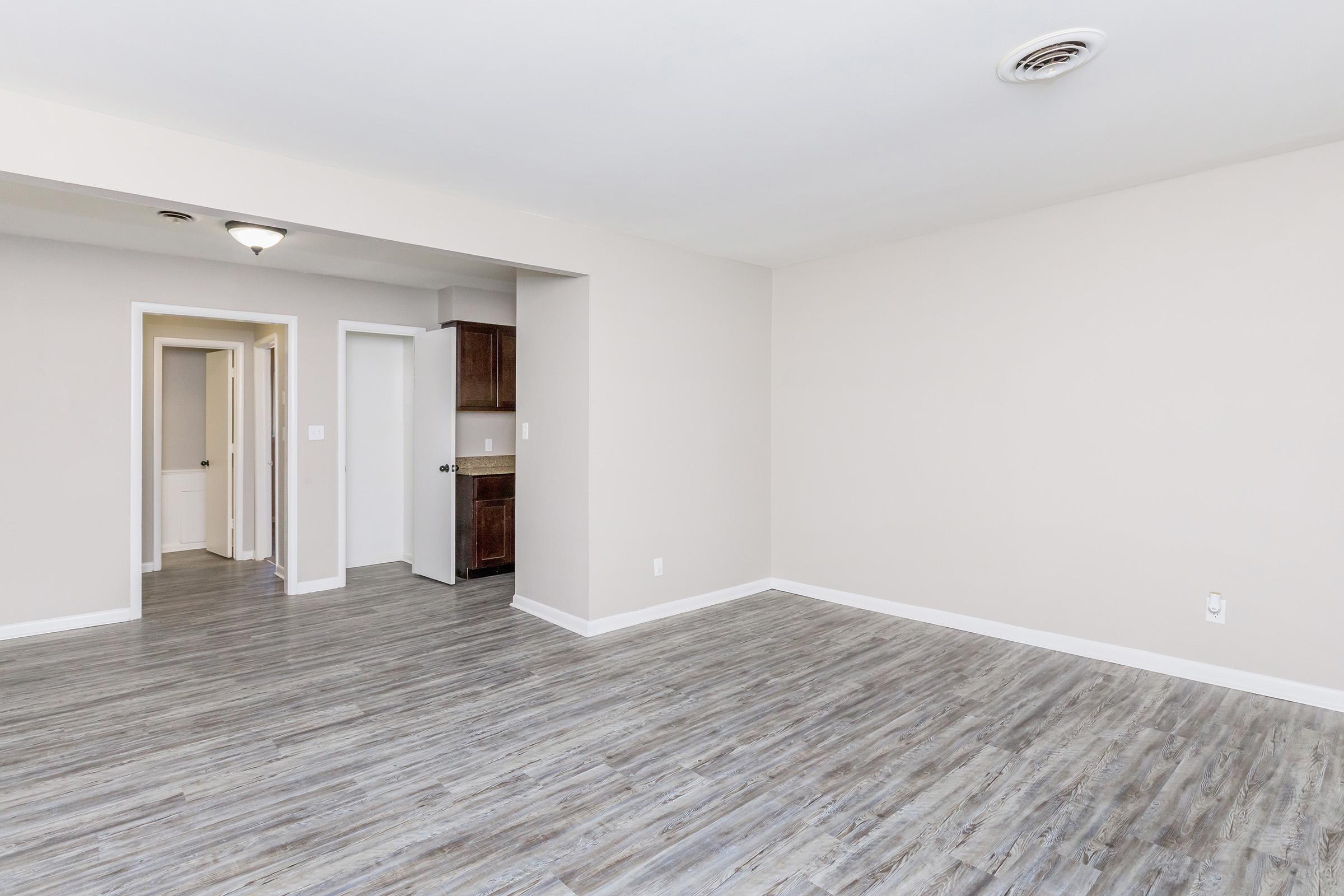 A spacious living area with light gray walls and hardwood-like flooring. There is a doorway leading to a hallway and a kitchen area with brown cabinetry in the background. The room is well-lit with a ceiling light fixture and has a minimalist, clean aesthetic.