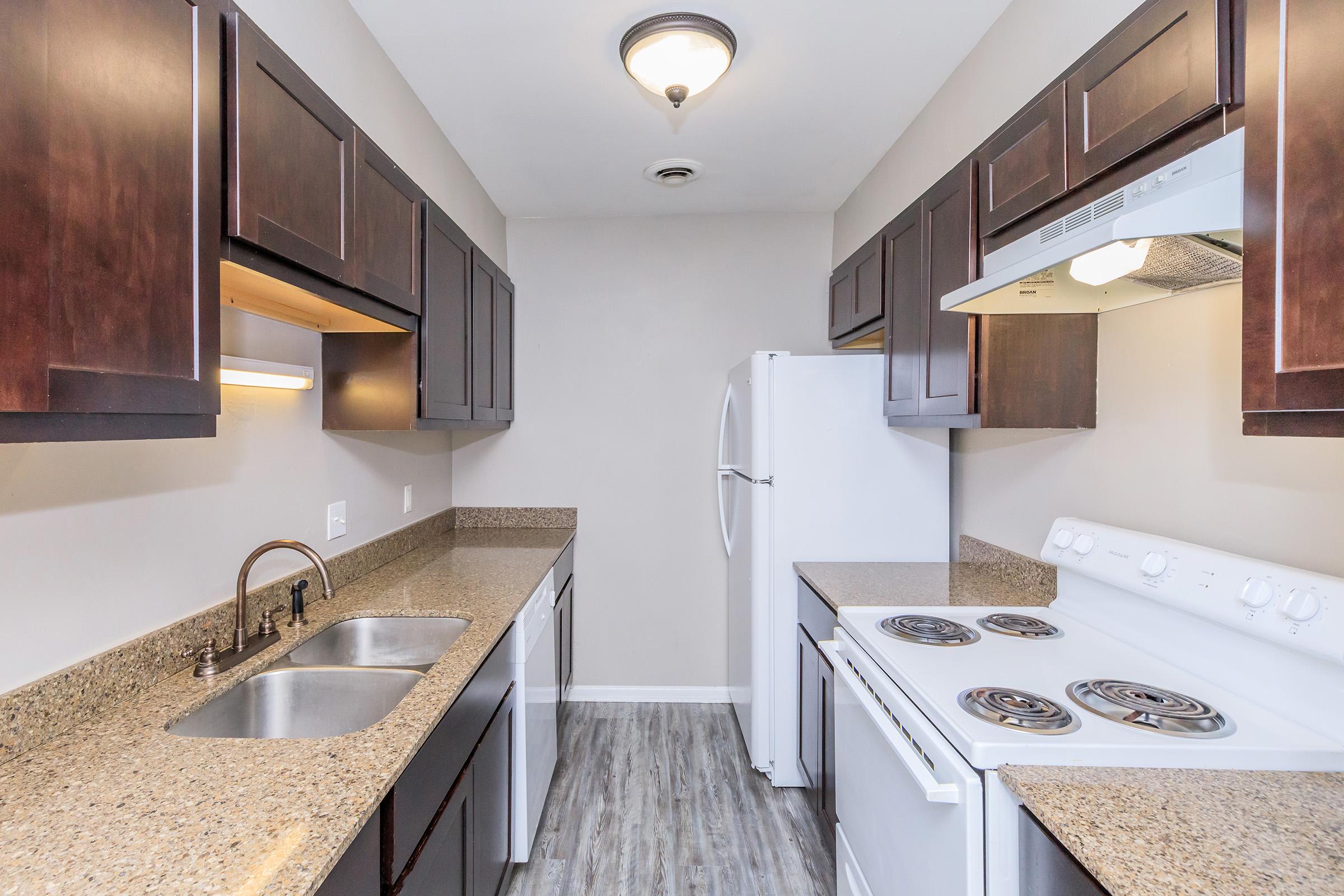 Modern kitchen featuring dark wood cabinets, dual sink, white appliances including a refrigerator and stove, and granite countertops. The space is well-lit with a ceiling light fixture and has a neutral-colored wall and floor.