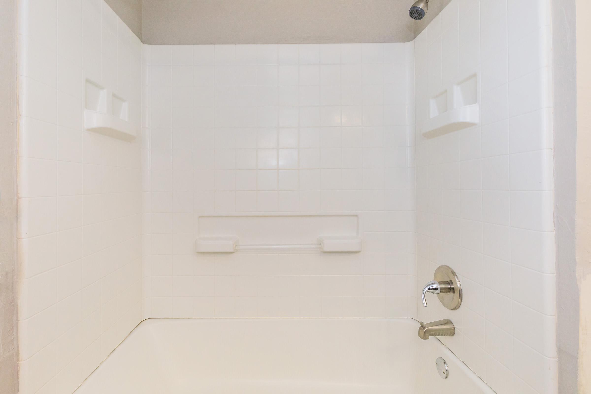 A clean, white tiled shower stall featuring two built-in shelves. The fixture includes a faucet and a showerhead mounted on the wall. The design is simple and modern, with a spacious tub base and minimal decorative elements. The overall appearance is bright and inviting.