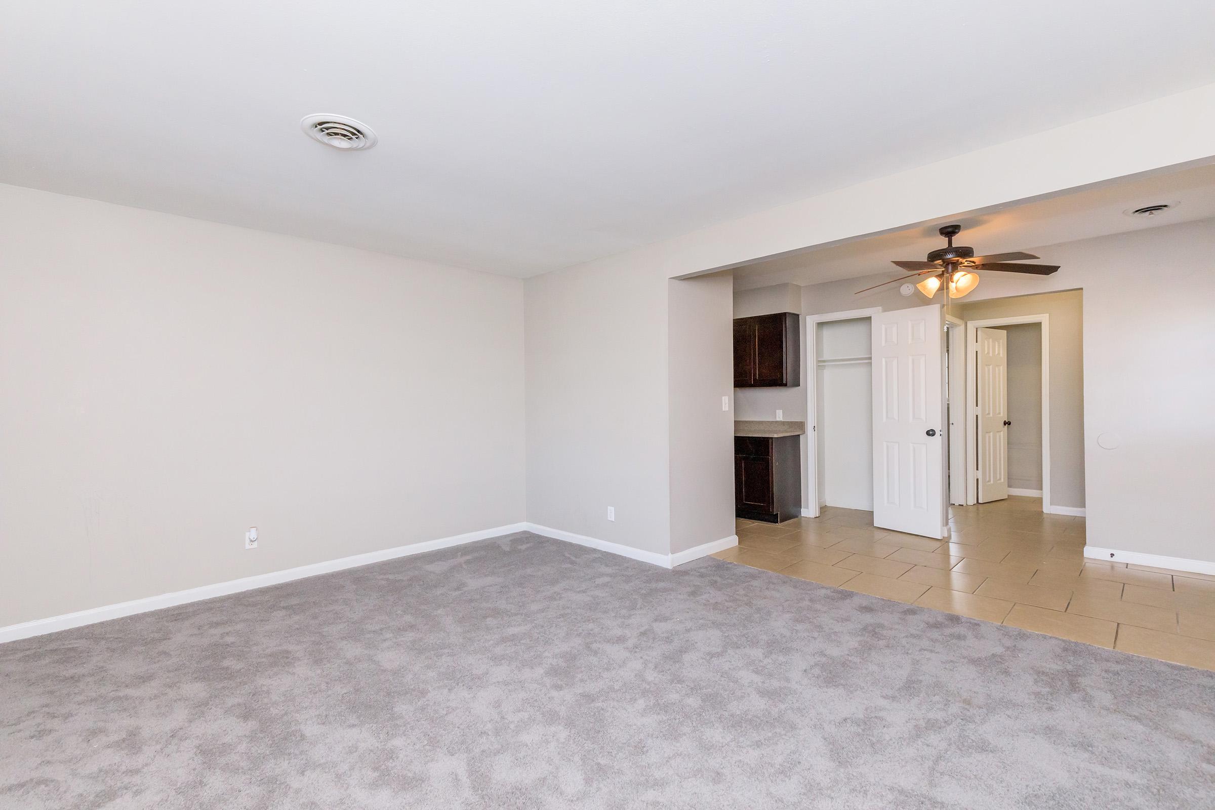 A spacious, empty living room with light gray carpet and neutral walls. A ceiling fan is visible, and a doorway leads to another room with tiled flooring and a small kitchen or dining area visible in the background. Natural light is present, creating a bright and open atmosphere.