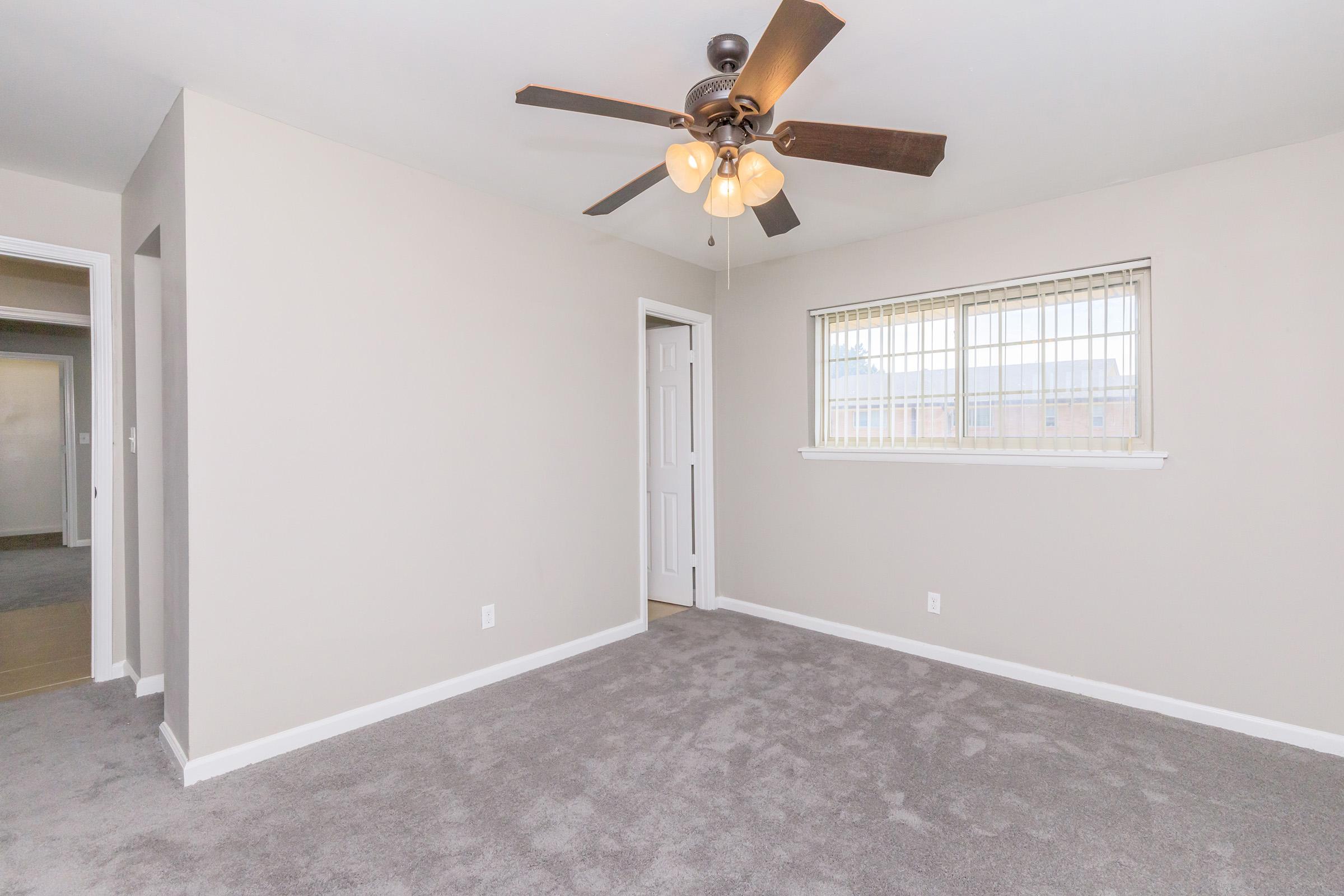 A well-lit, empty room with light gray walls and carpeting. It features a ceiling fan with four light bulbs and a window with blinds. A door to the left leads to another room, while a doorway on the right is partially visible. The space is clean and modern, suitable for various furnishings.