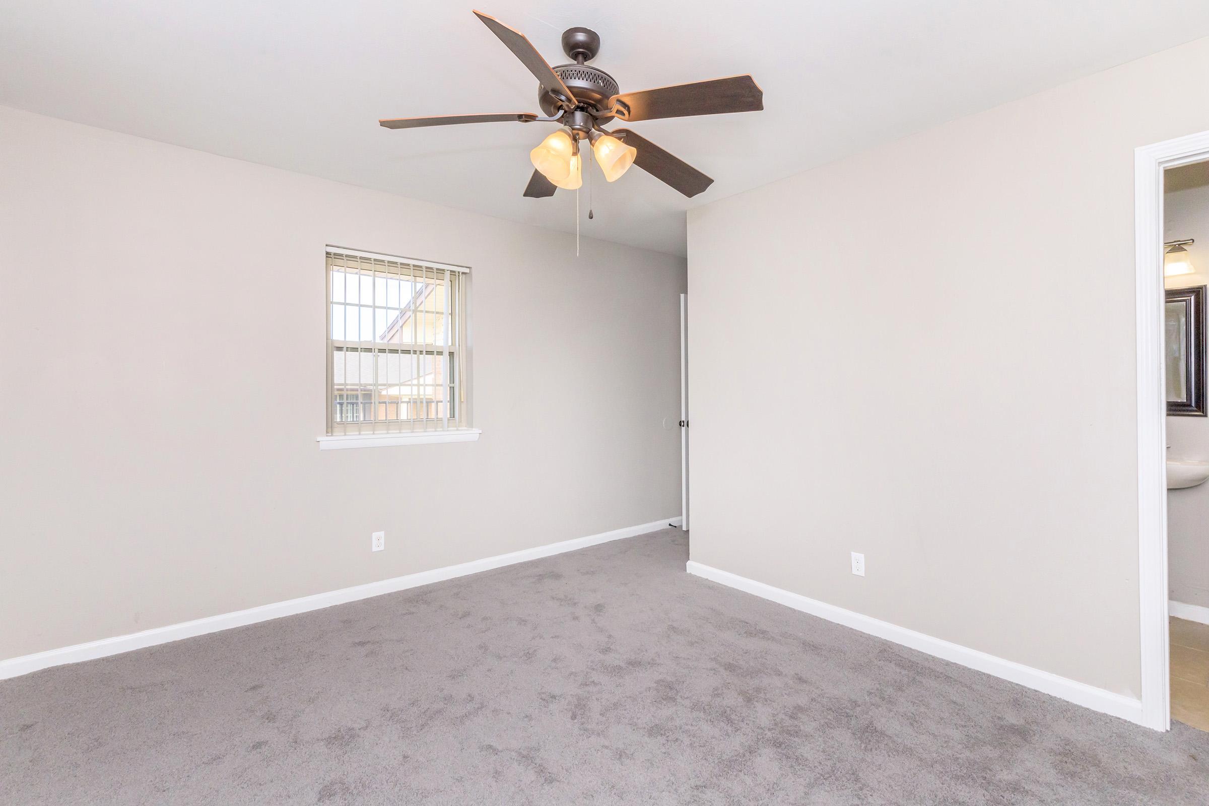 A spacious, lightly colored room featuring beige walls and a plush carpet. A ceiling fan with three light fixtures hangs from the ceiling, providing additional light. A window is located on one wall, allowing natural light to enter, and a door leads to another area of the house.