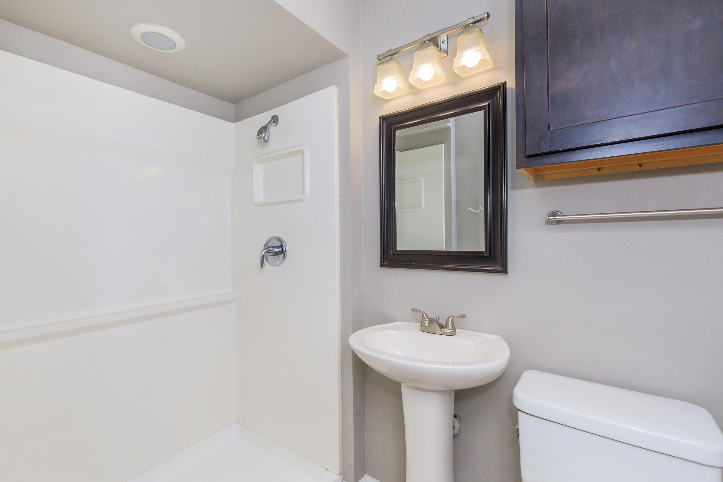 A clean and modern bathroom featuring a shower with white walls, a pedestal sink, a toilet, and a mirror with a dark frame. The lighting fixtures above the mirror provide bright illumination, enhancing the overall aesthetic of the space.