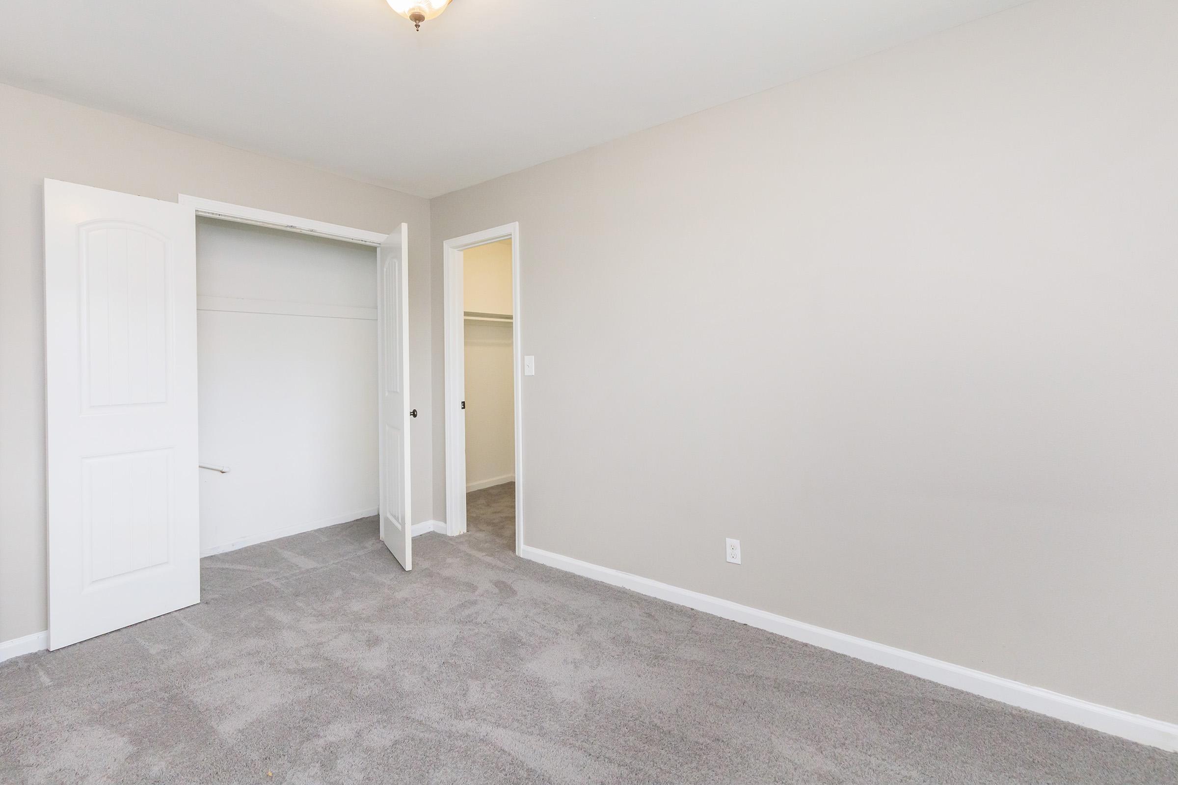A vacant room with light gray carpet and neutral walls. There are two open doors: one leading to a closet and another to a small adjoining room. The ceiling has a simple light fixture. The space appears bright and uncluttered, ideal for potential furnishings or decor.