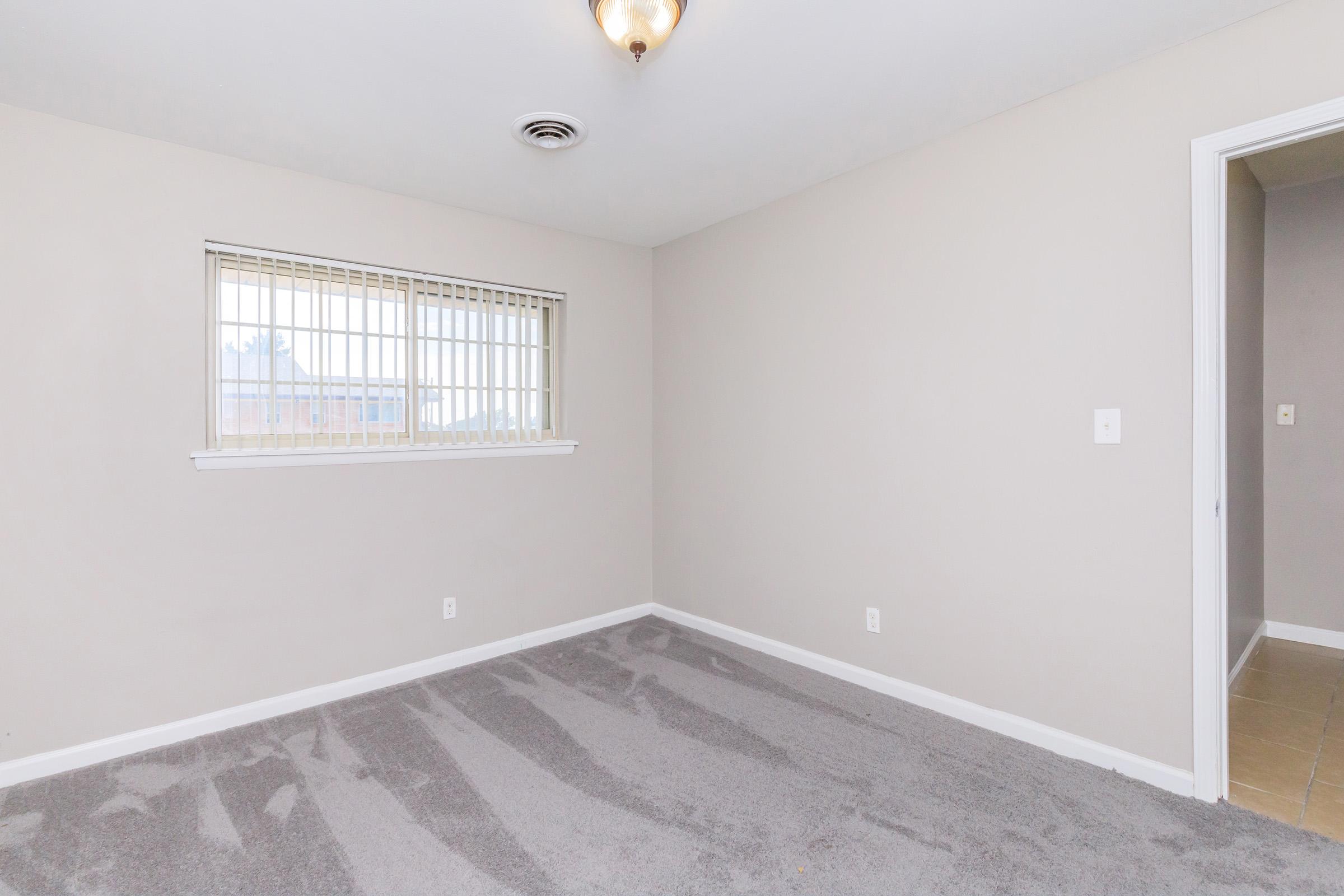 Empty room with light beige walls, a window with blinds, and new carpeting. The room features a ceiling light fixture and a doorway leading to another space. The floor is partially tiled near the entrance. Natural light comes in through the window, creating a bright and airy atmosphere.