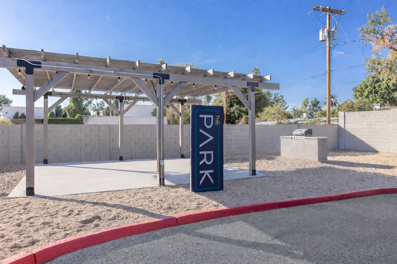 A sheltered outdoor area at a park featuring a large sign that reads "THE PARK." The space is surrounded by gravel and has a barbecue grill in the background. There are utility poles and blue skies, indicating a clear day.
