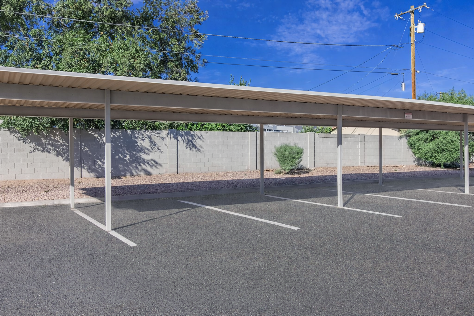 A carport structure with several empty parking spaces beneath it. The background features a gray wall and some low shrubs, with a blue sky and scattered clouds above. Utility poles are visible in the distance. The area appears clean and well-maintained.