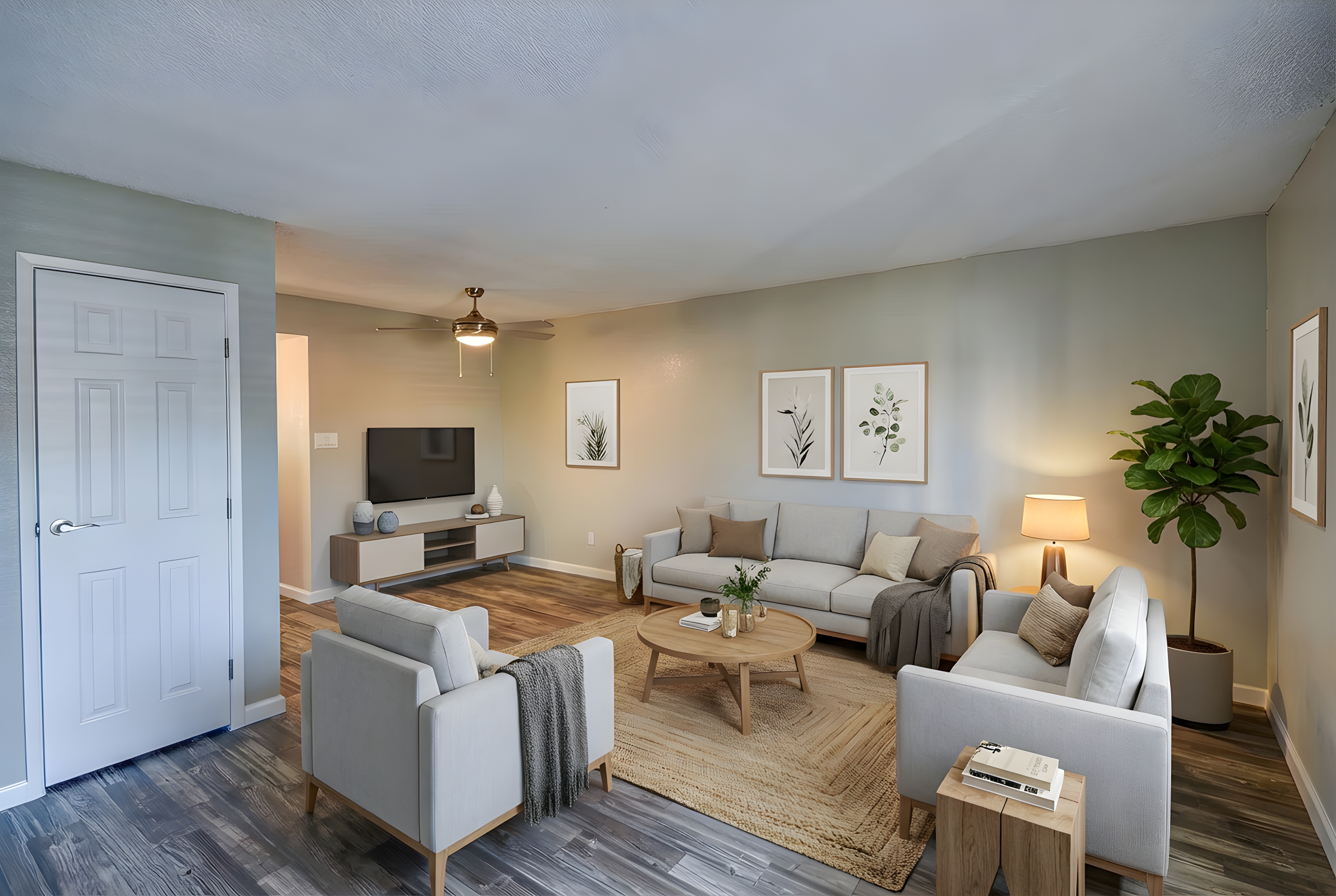 A modern living room featuring a light gray color palette, a comfortable sofa, and two armchairs. A round coffee table sits in the center on a textured area rug. There are framed botanical prints on the wall, a floor lamp beside the sofa, and a potted plant in the corner. A TV is mounted on the wall across from the seating area.