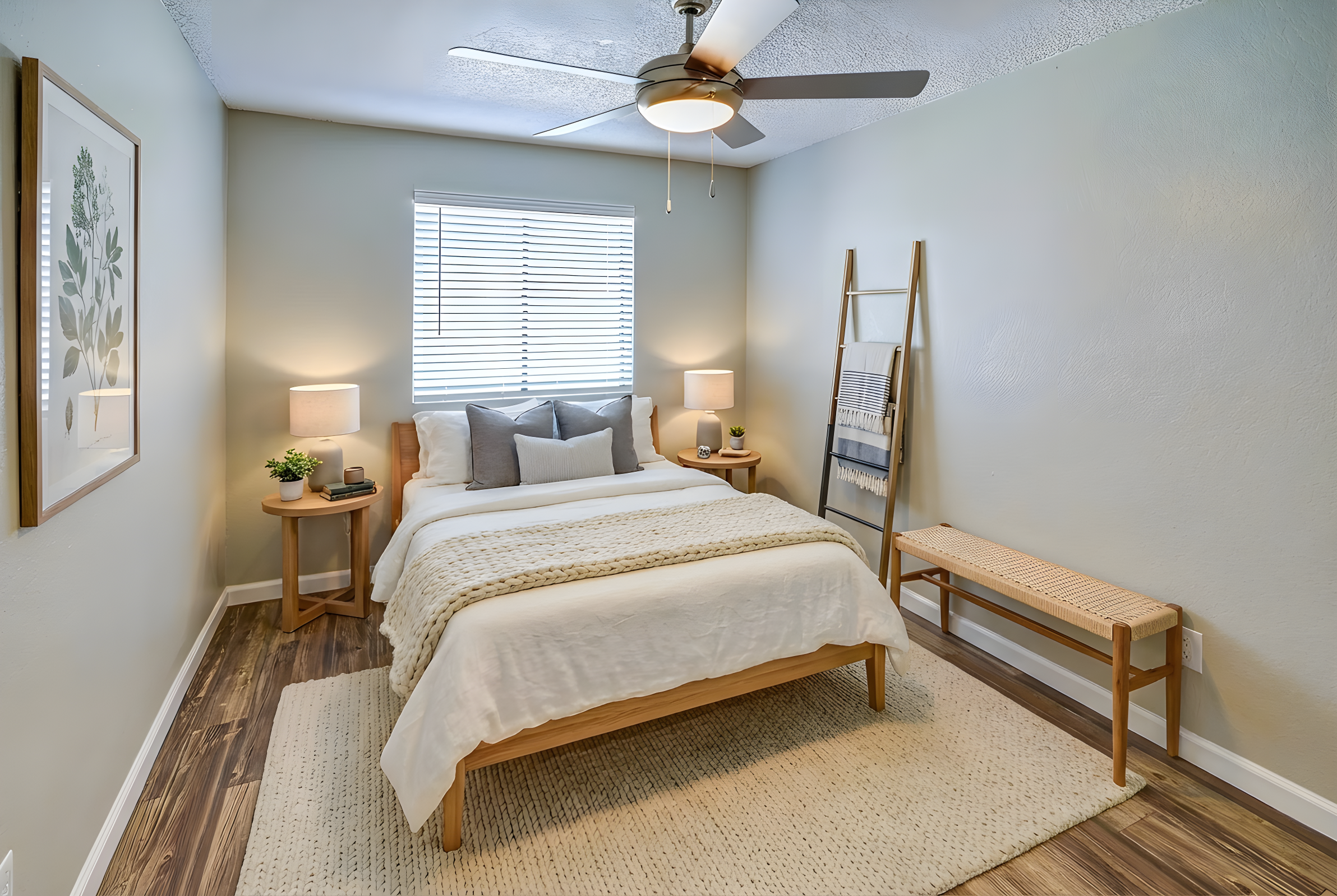 A cozy, modern bedroom featuring a queen-sized bed with neutral bedding, two bedside tables with lamps, and a decorative ladder holding towels. The room has a light green wall, a window with blinds, and a woven rug on the wooden floor, creating a warm and inviting atmosphere.