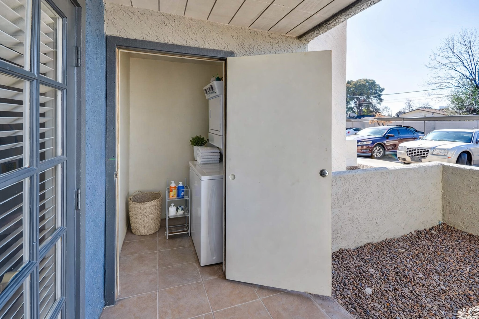 A partially open door reveals a small laundry area with a washing machine and storage items, alongside a woven basket. Outside the door, a gravel area can be seen, and parked vehicles are visible in the background under a clear blue sky.