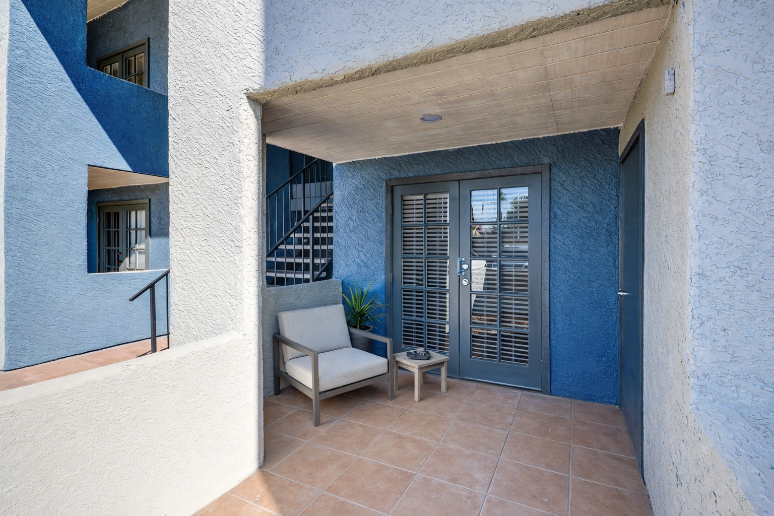 A cozy entrance area featuring a pair of stylish chairs and a small table. The walls are painted in shades of blue and light gray, and a potted plant adds a touch of greenery. A staircase is visible in the background, leading to the upper level of the building. Light spills in from the door, creating a welcoming atmosphere.