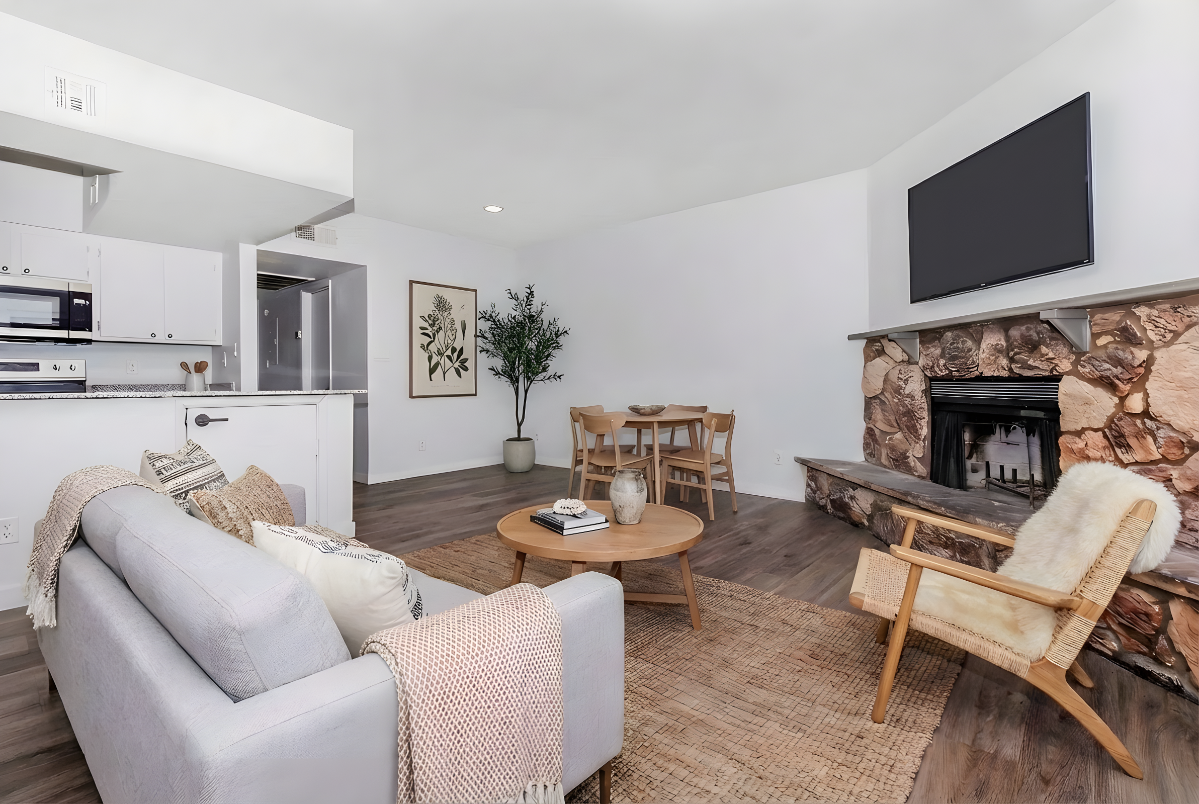 A modern living room featuring a light gray sofa with a blanket, a wooden coffee table, and a cozy chair. There is a stone fireplace with a TV above, hardwood flooring, and a decorative plant near a dining area with a circular table and chairs. The walls are painted white for a bright, airy feel.