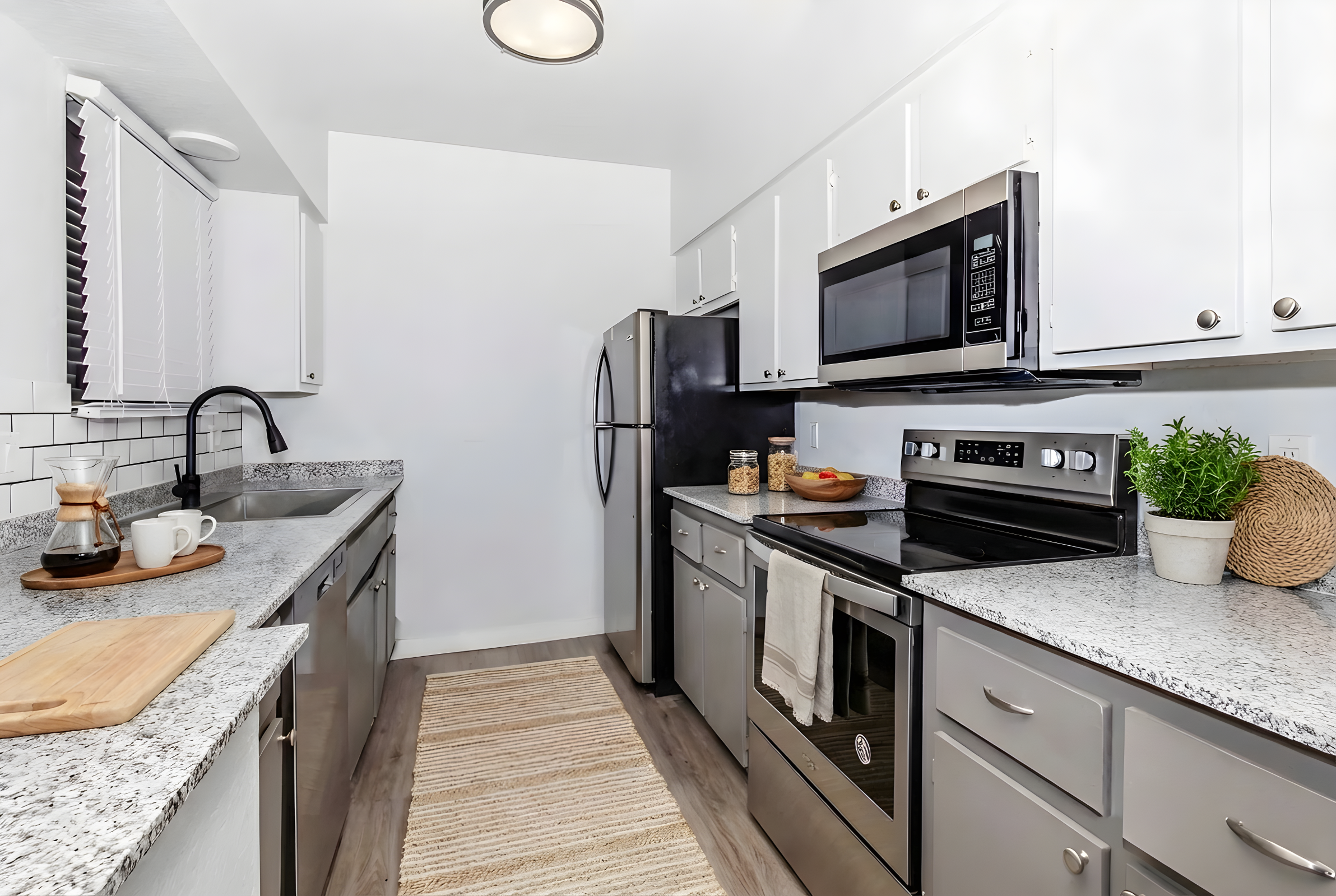 Modern kitchen featuring gray cabinets, stainless steel appliances including a refrigerator and microwave, a granite countertop, and a coffee maker on a wooden cutting board. Natural light filters in through a window, illuminating decorative plants and a neutral-toned rug on the floor.