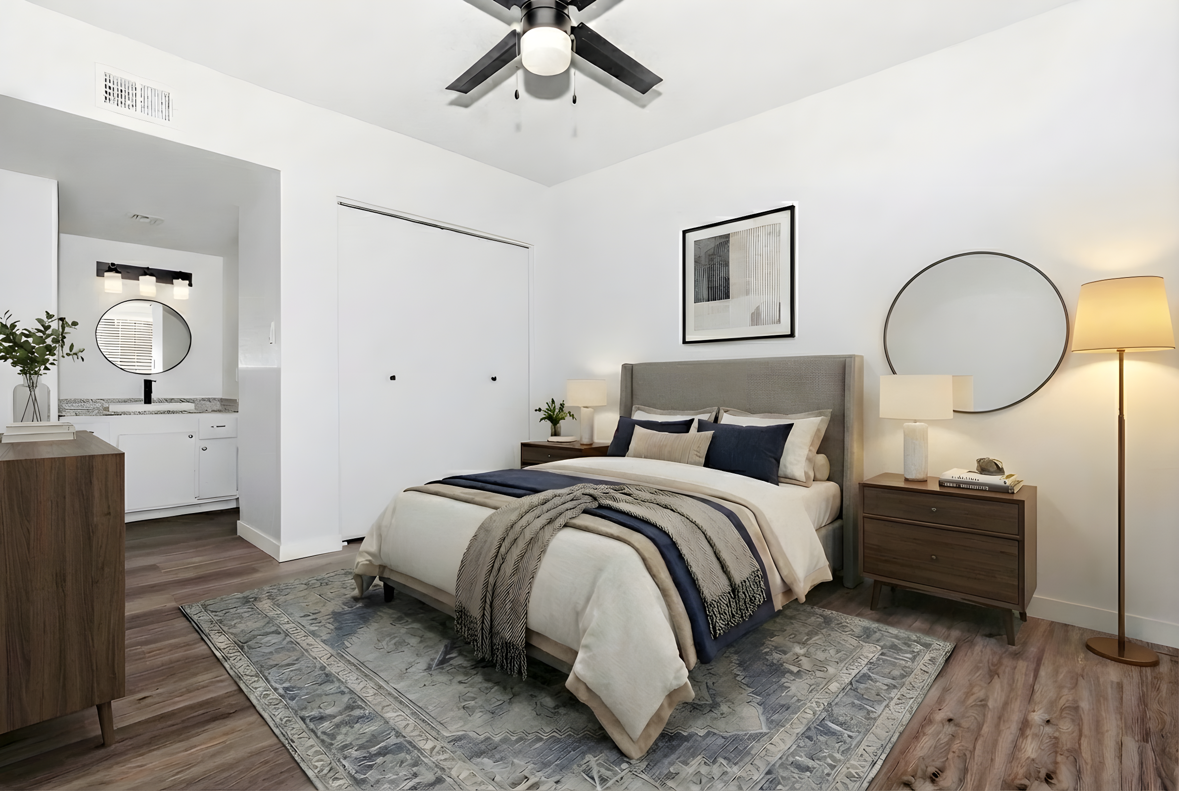 A modern bedroom featuring a queen-sized bed with dark blue and beige bedding, complemented by a decorative throw. There are two wooden nightstands with lamps, a round mirror on the wall, and a light-colored area rug on the floor. A bathroom is visible through an open door in the background.