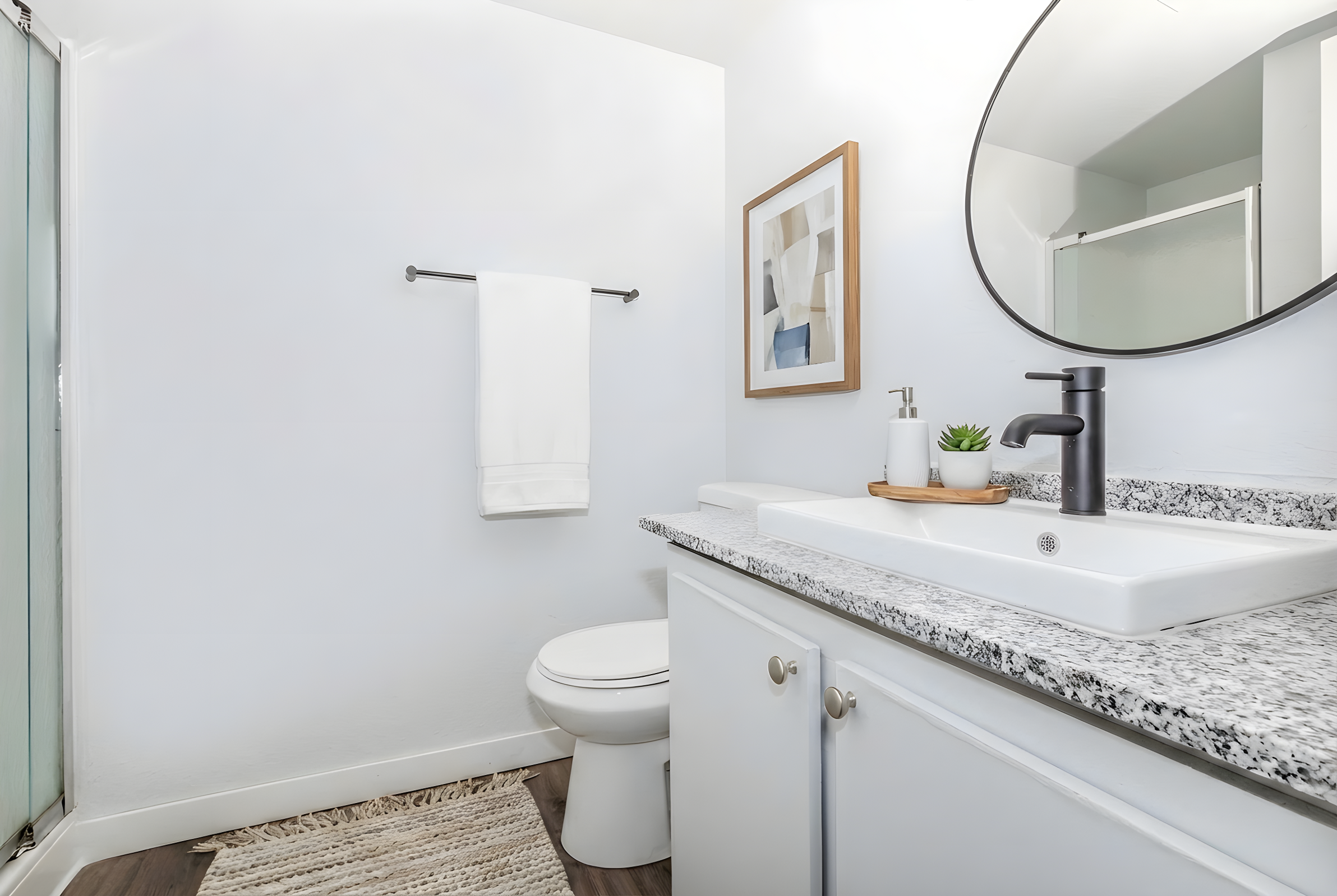 Bright and modern bathroom featuring a white toilet and a sleek countertop with a rectangular sink. A white towel hangs on a towel rack, and a small plant on a wooden tray adds a touch of greenery. The walls are painted light, enhancing the spacious feel, and there is a glass shower enclosure visible in the background.
