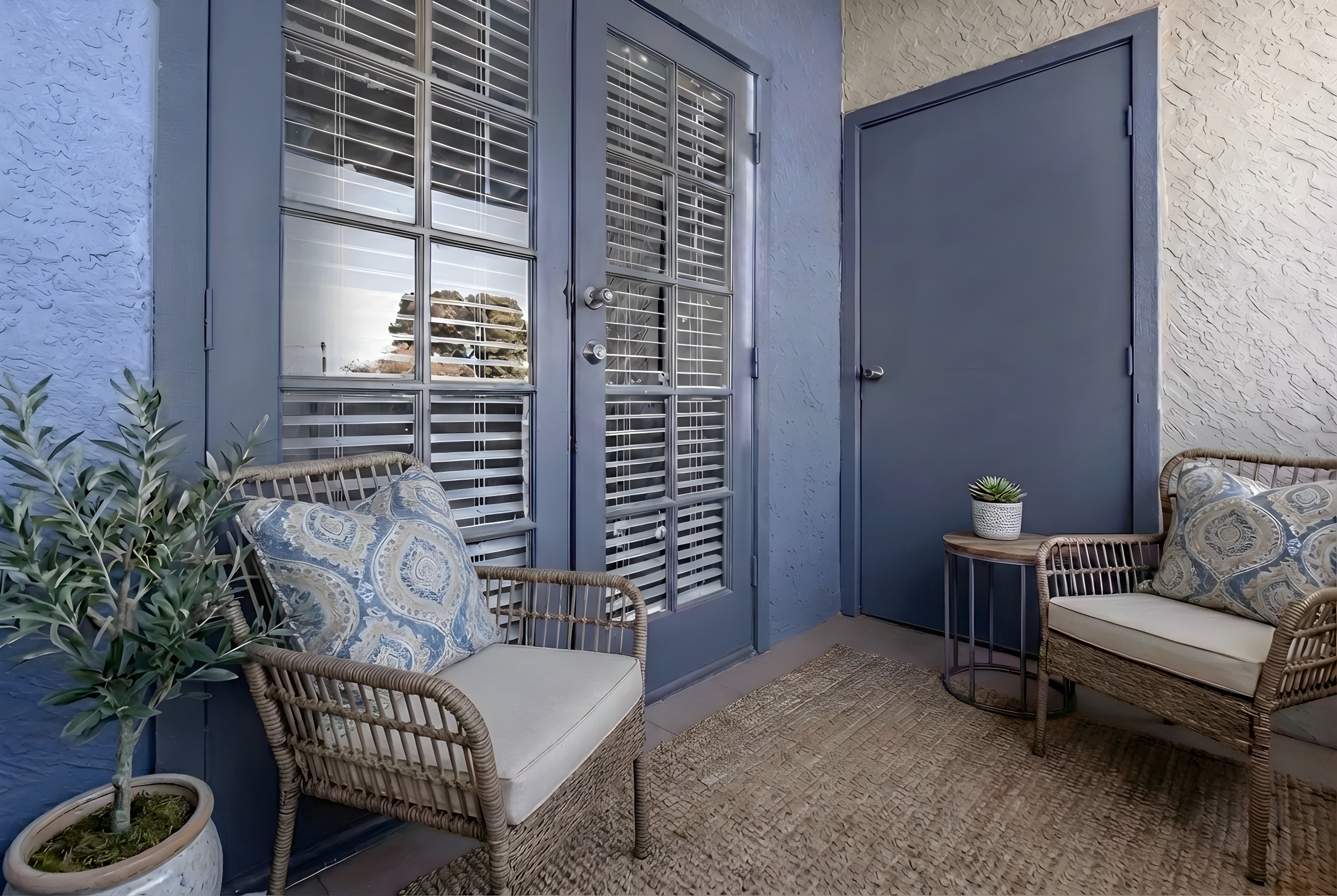 A cozy outdoor seating area featuring two wicker chairs with patterned cushions, a small potted plant on a table, a textured rug, and a door with shutters, all against a blue and beige wall backdrop. The scene offers a welcoming atmosphere for relaxation.