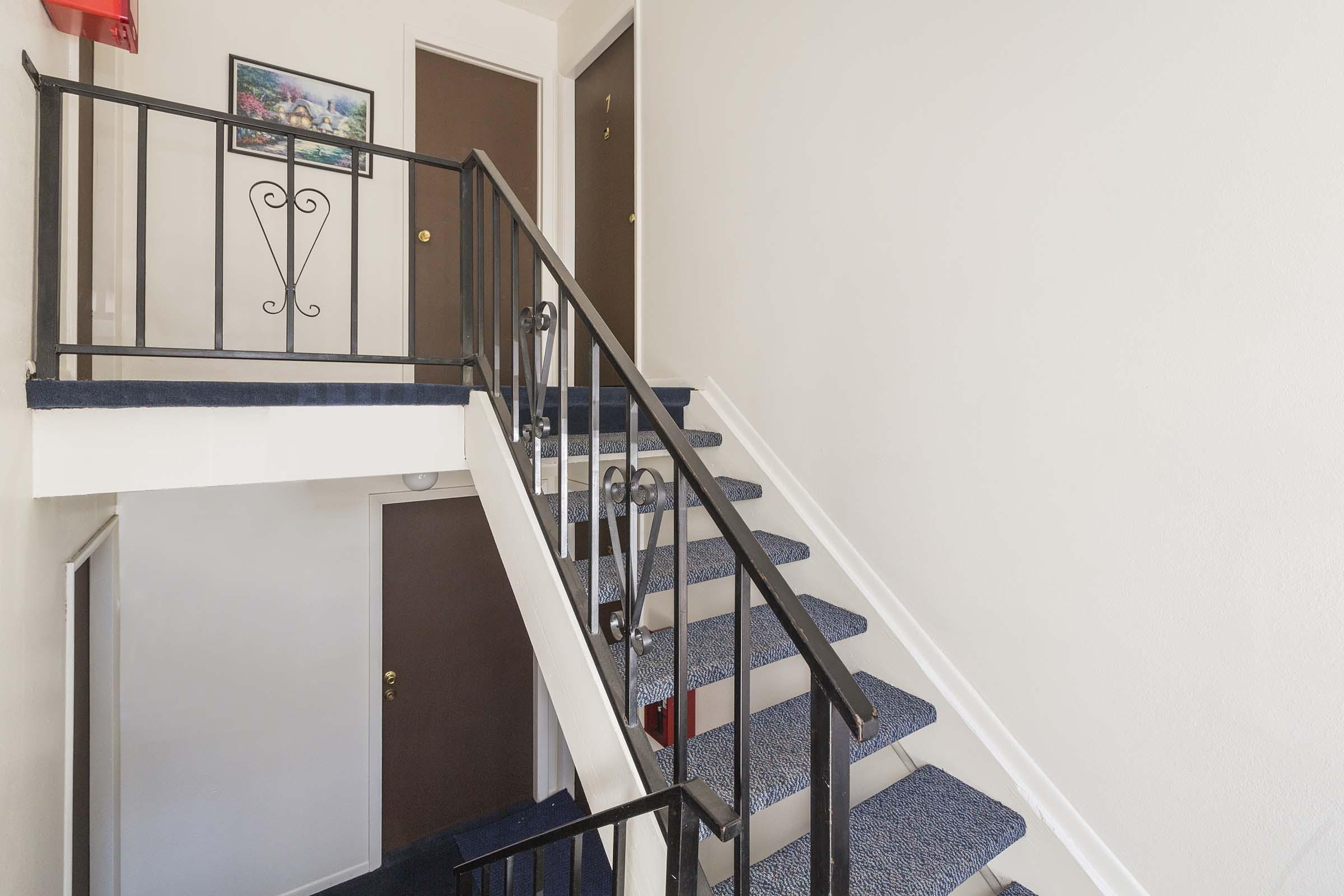 Interior view of a staircase in a building, featuring a dark blue carpet and a black railing. A wall-mounted artwork is visible in the background, along with two closed doors, one with a small round doorknob and the other with a standard door handle. The walls are painted a light color.