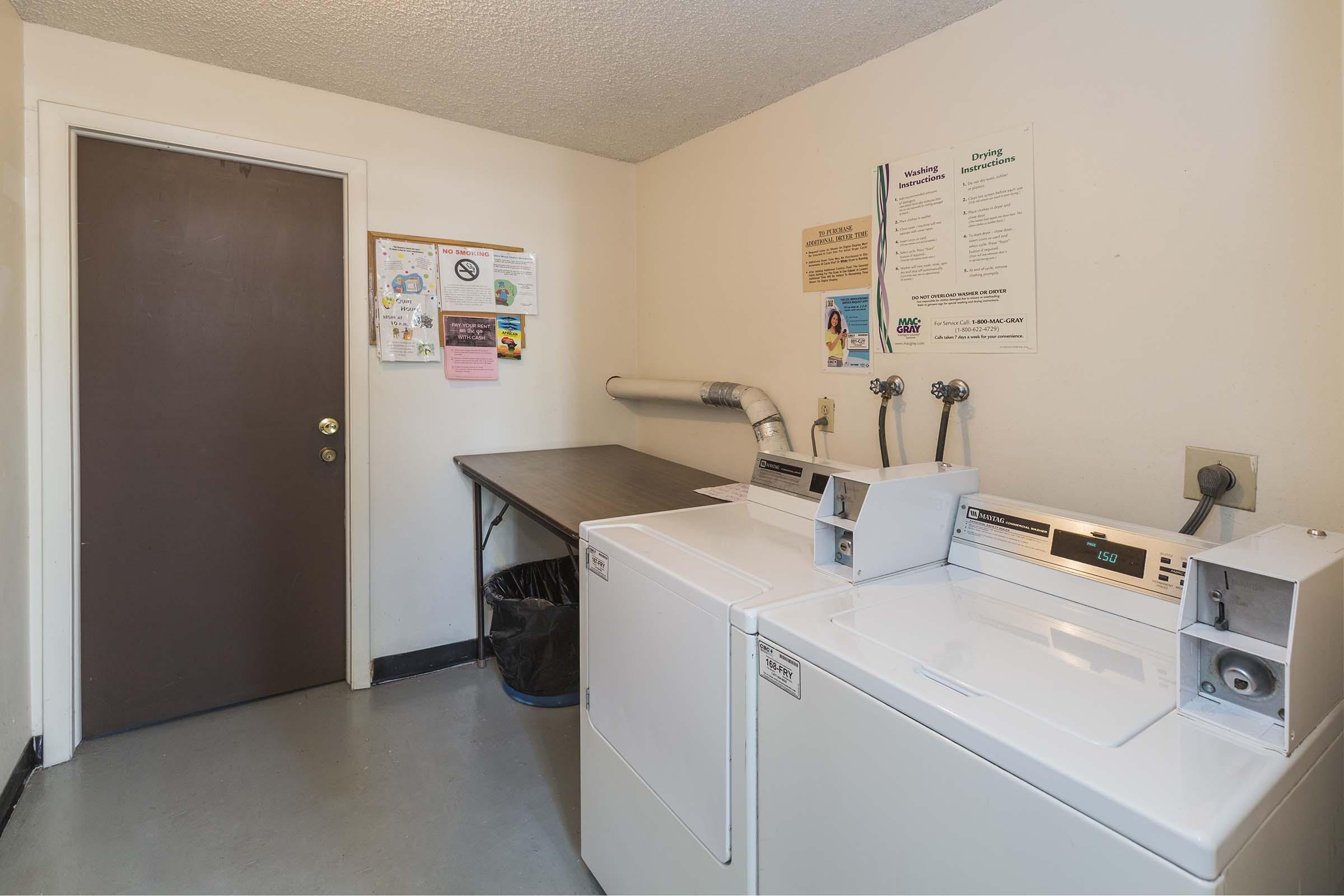A clean laundry room featuring two white washing machines side by side, a metal folding table, and a large trash bin. The walls are decorated with notices and instructions. A plain door is visible in the background, providing access to the room.