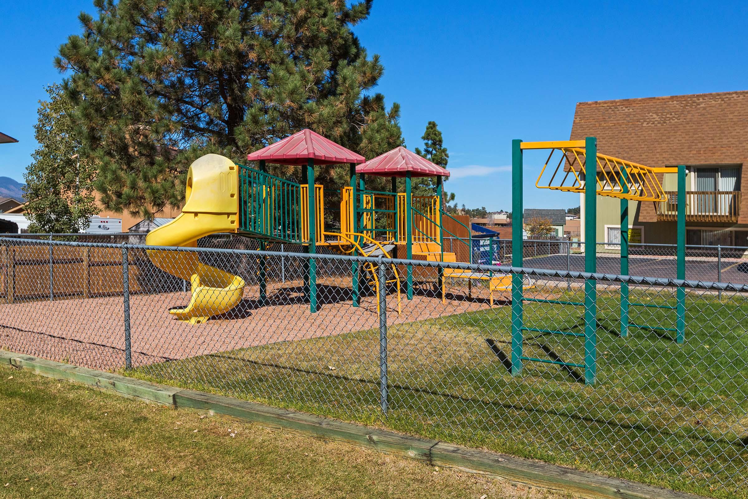 A colorful playground featuring a yellow slide, climbing structures, and monkey bars, enclosed by a chain-link fence. In the background, there are trees and a building with a brown roof under a clear blue sky.