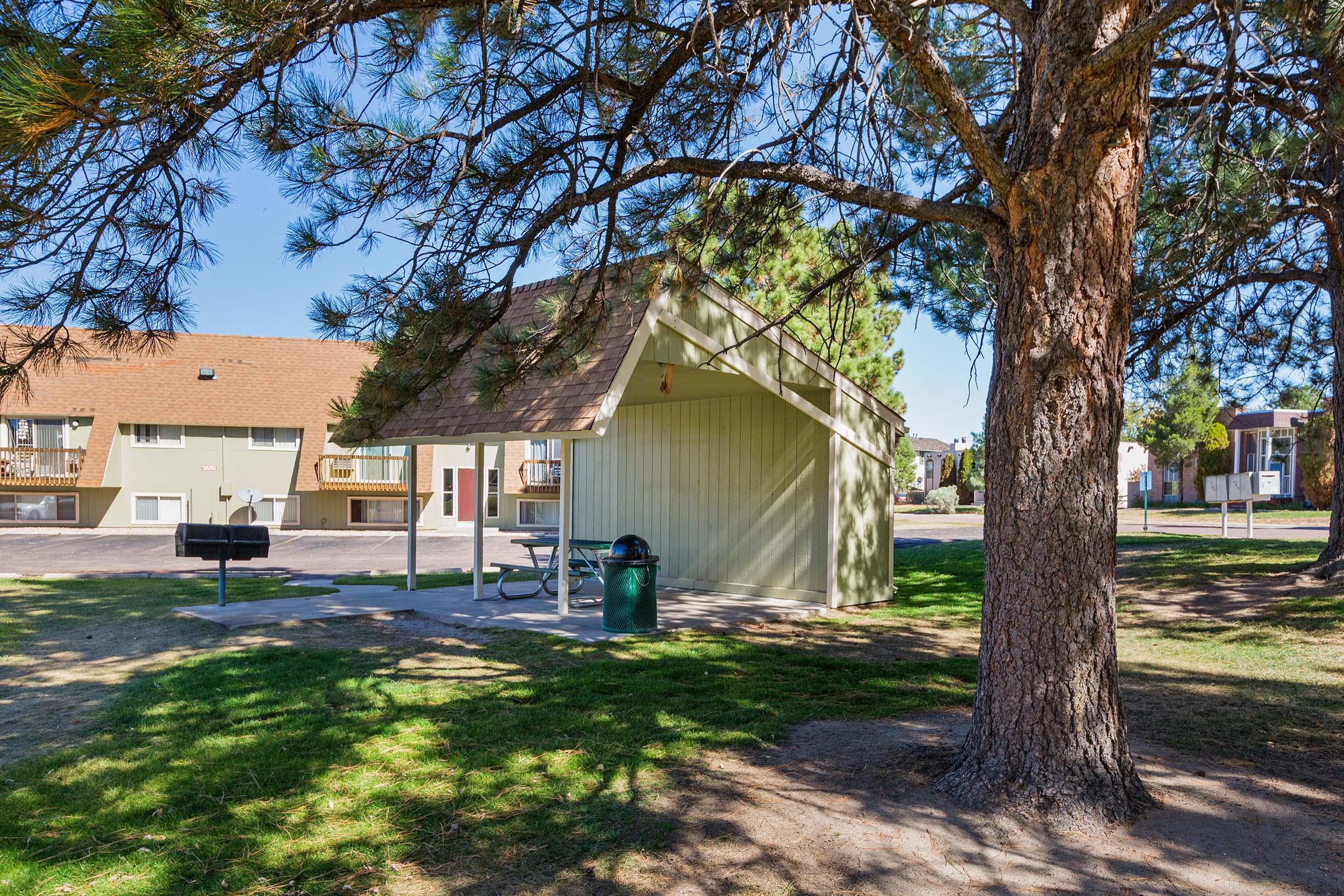 A shaded picnic area surrounded by trees, featuring a small shelter with a grill and a trash can. In the background, several residential buildings are visible under a clear blue sky. The scene captures a peaceful outdoor setting suitable for gatherings.
