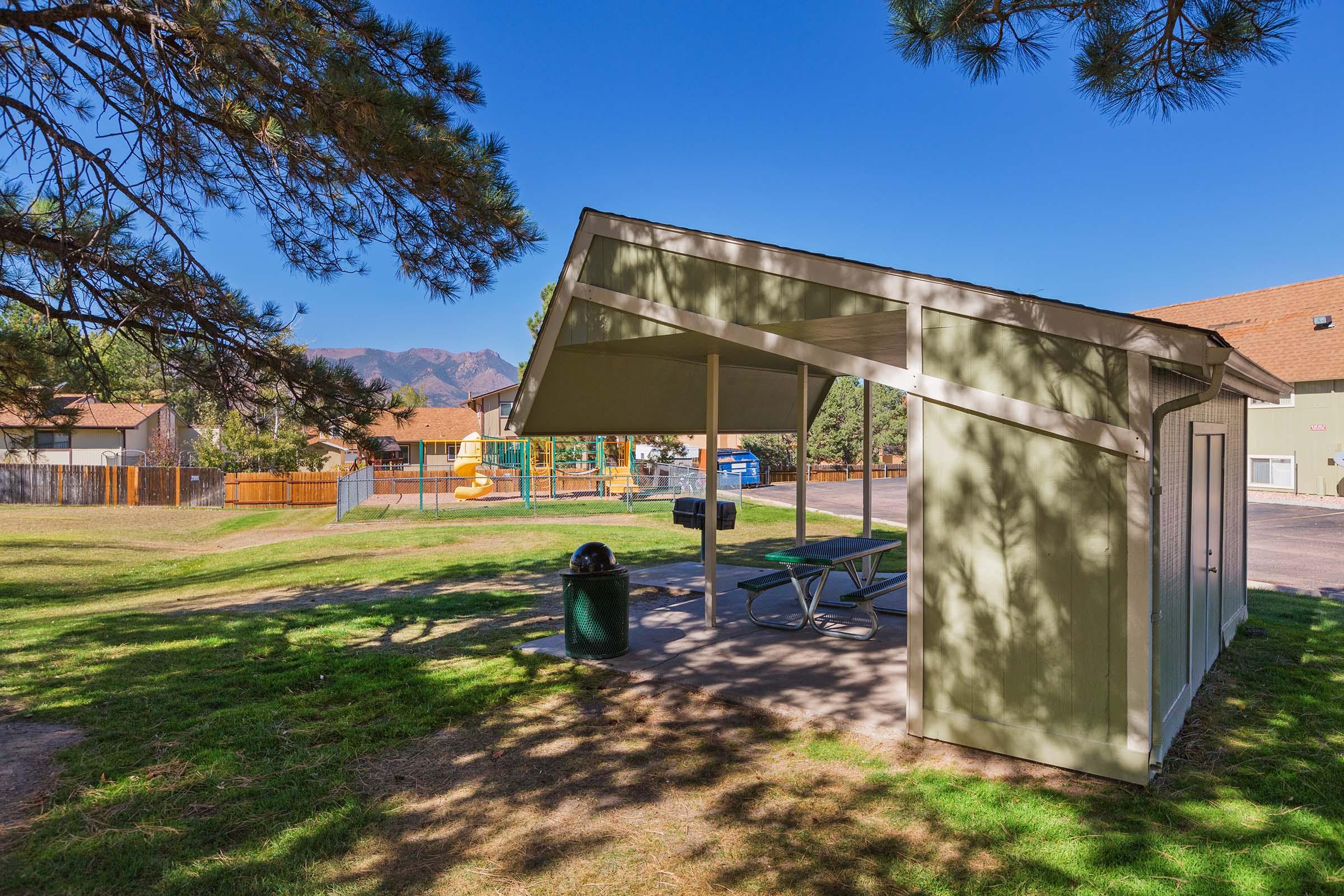 A shaded picnic shelter in a park, surrounded by grass and trees. In the background, there is a playground with colorful equipment and a fenced area. The sky is clear and blue, with mountains visible in the distance.
