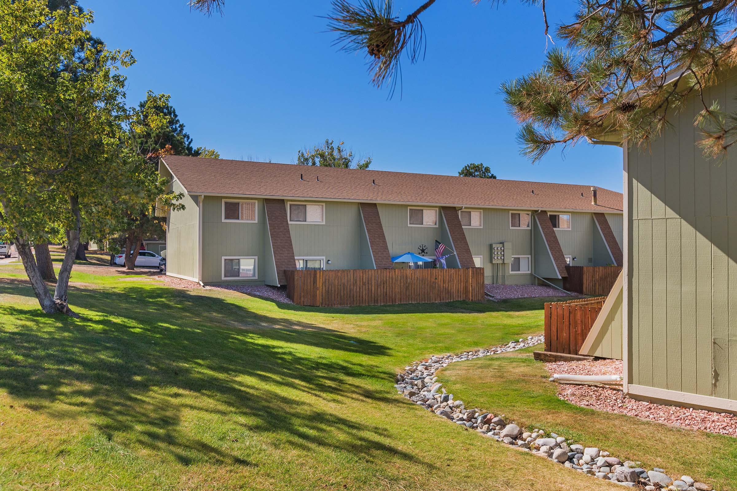 A well-maintained apartment complex surrounded by green lawns and trees under a clear blue sky. The building features a series of balconies, and some units have American flags displayed. A stone path winds through the grass, adding to the inviting atmosphere.
