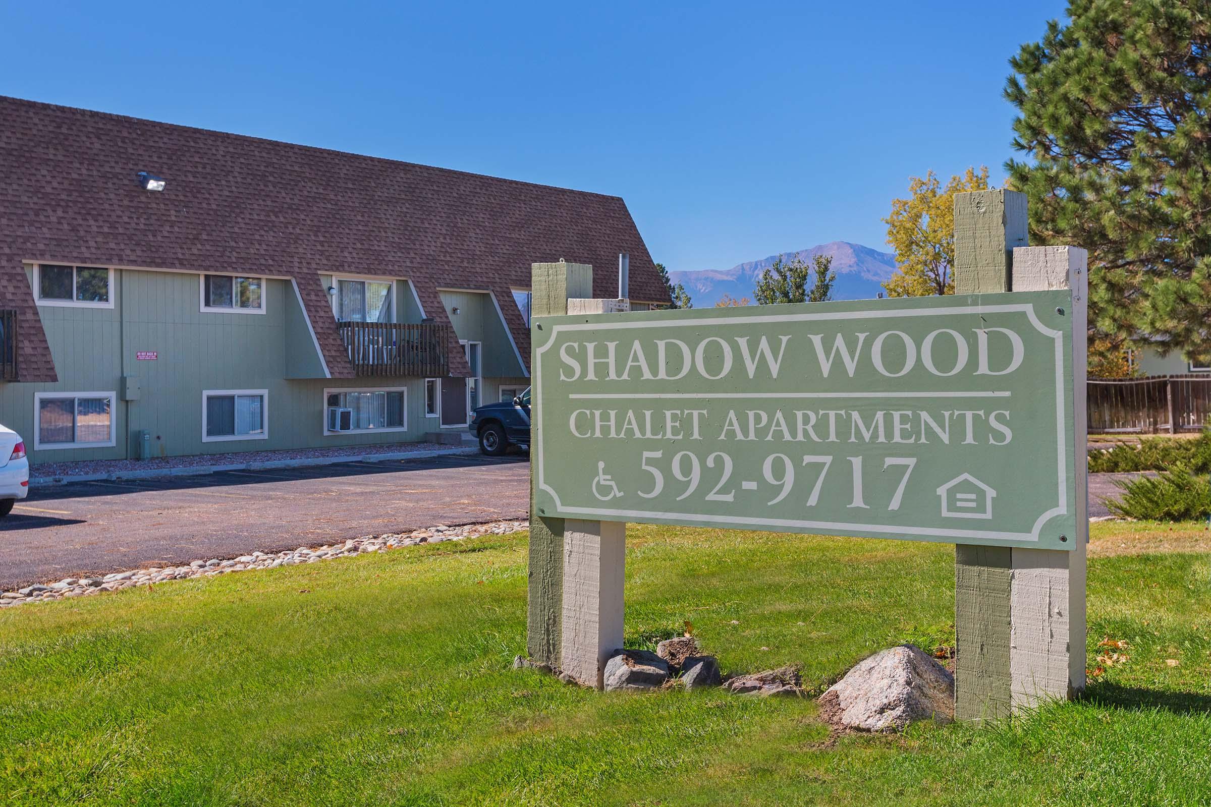 Sign for Shadow Wood Chalet Apartments, featuring the name and contact number (592-9717). The background shows a multi-unit building with a sloped roof and a clear blue sky, surrounded by green grass and a few trees.