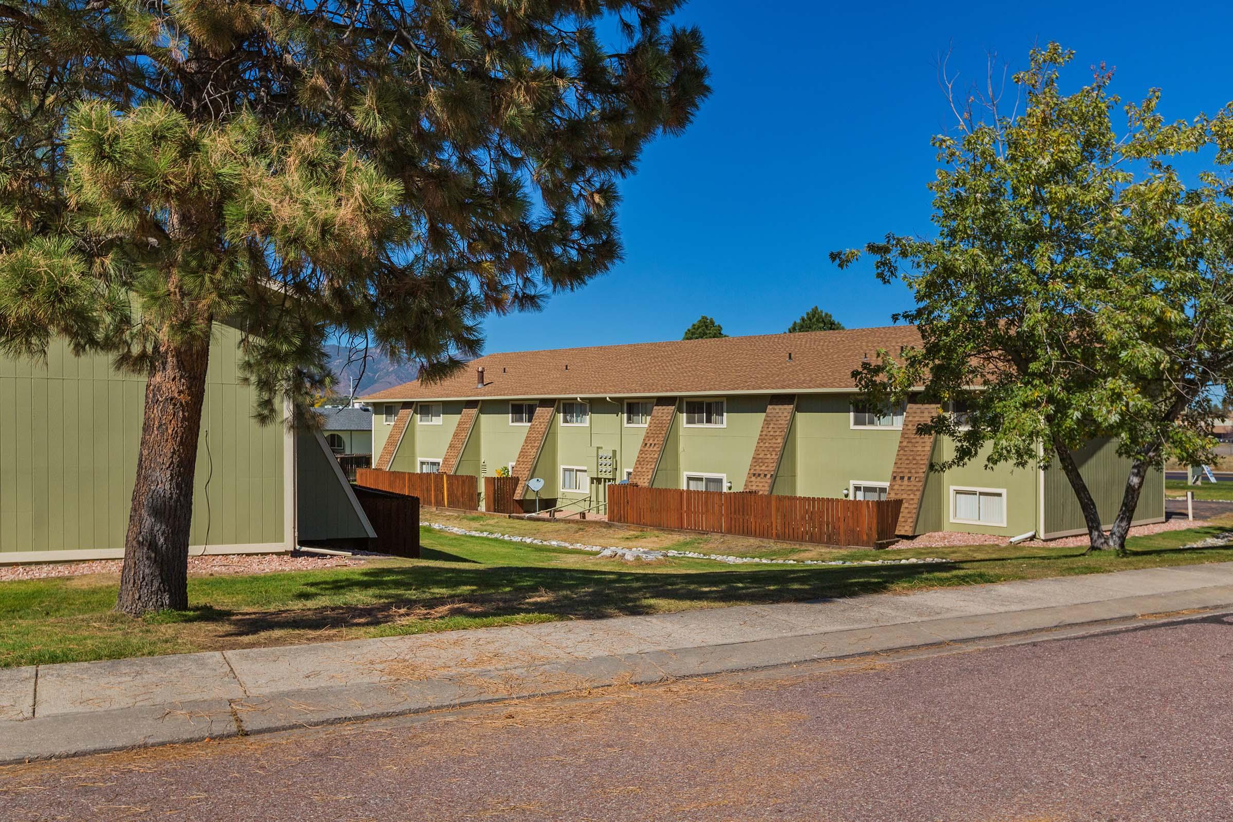 Two-story green apartment buildings with brown roofs, situated on a grassy area. There are trees on either side and a pathway running in front. The sky is clear and blue, indicating a sunny day.