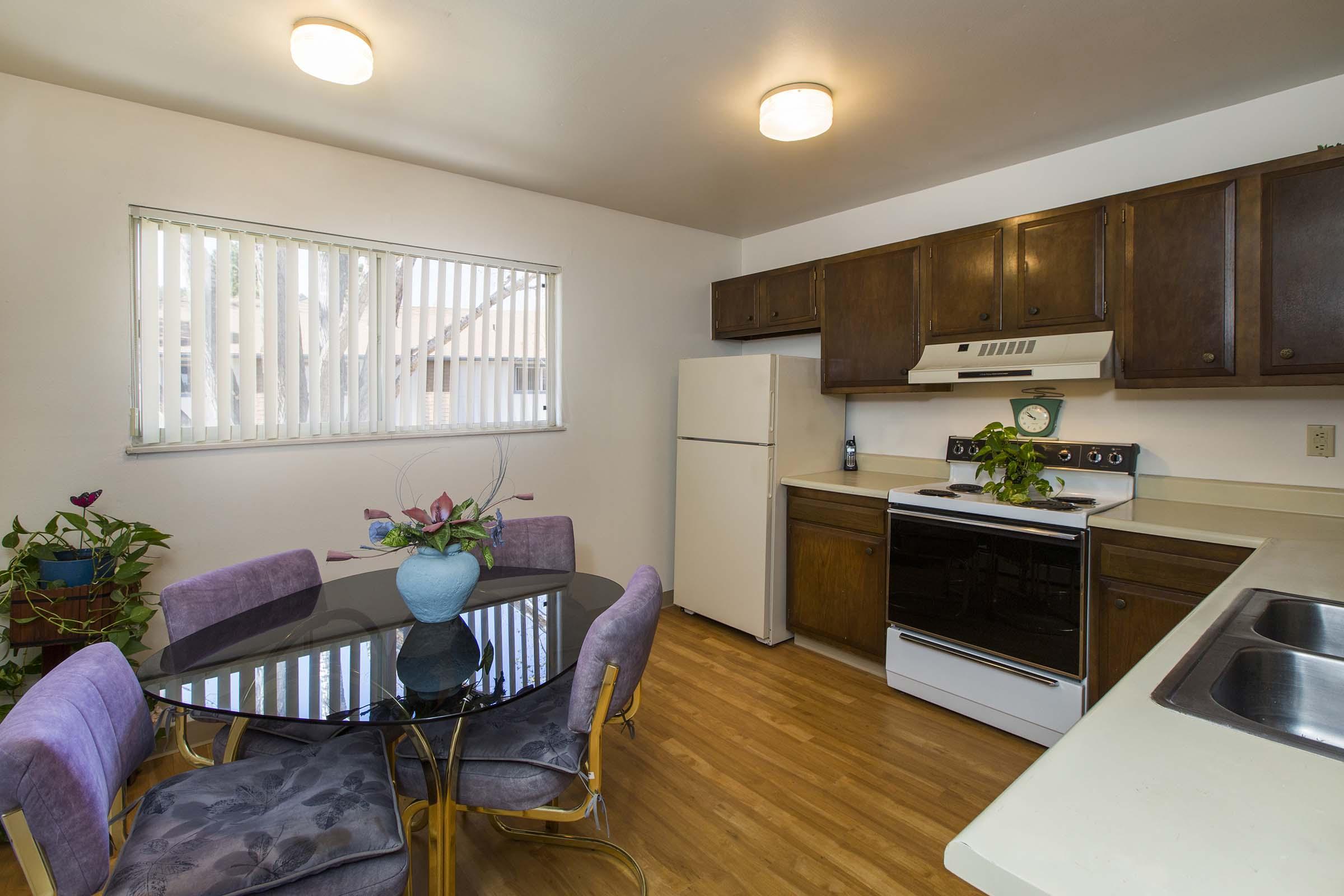 A bright kitchen with wooden cabinets, a white refrigerator, and an oven. There's a round glass dining table surrounded by purple chairs, and a vase with plants at the center. Sunlight filters through a window with horizontal blinds, illuminating the warm wooden floor.