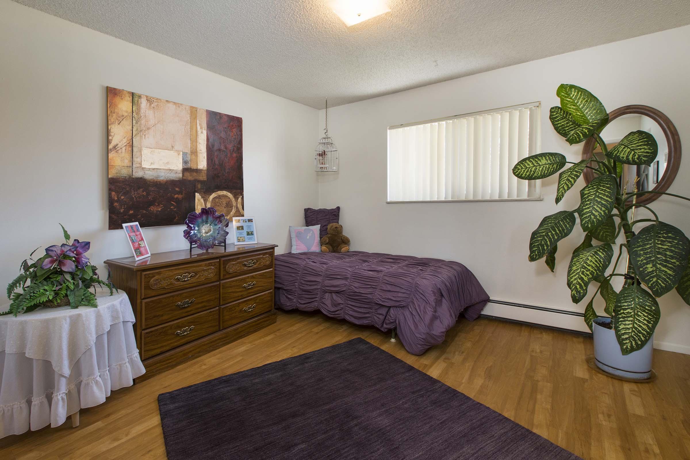 A cozy bedroom featuring a bed with a deep purple comforter, a decorative plant in the corner, a wooden dresser, a small table with flower arrangements, and a piece of artwork on the wall. Natural light comes in through a window covered with blinds.