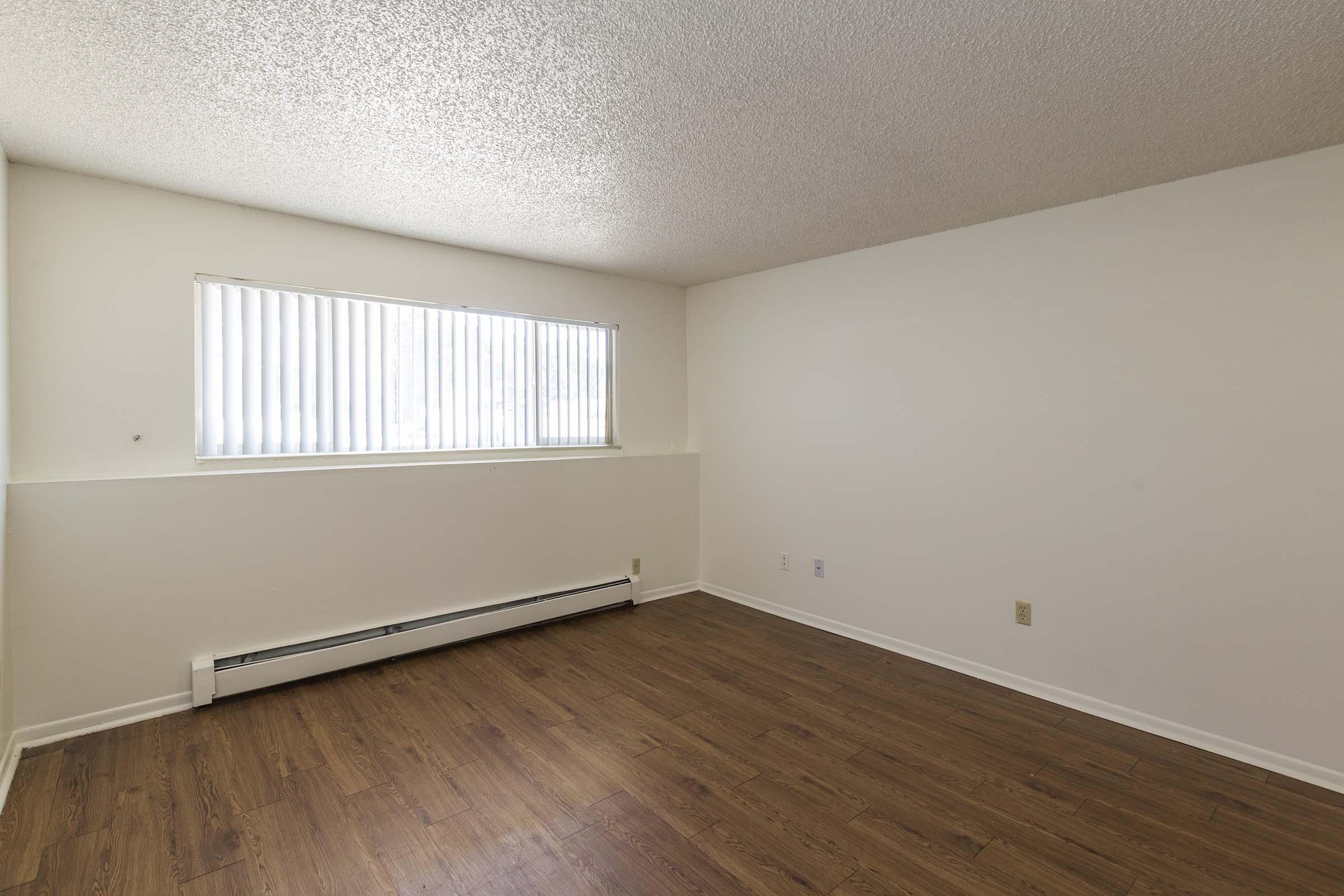 A sparsely furnished room with light-colored walls and a large window featuring vertical blinds. The floor is covered with a medium brown laminate and there is a baseboard heater along the wall. The space appears bright and empty, suitable for customization or staging.