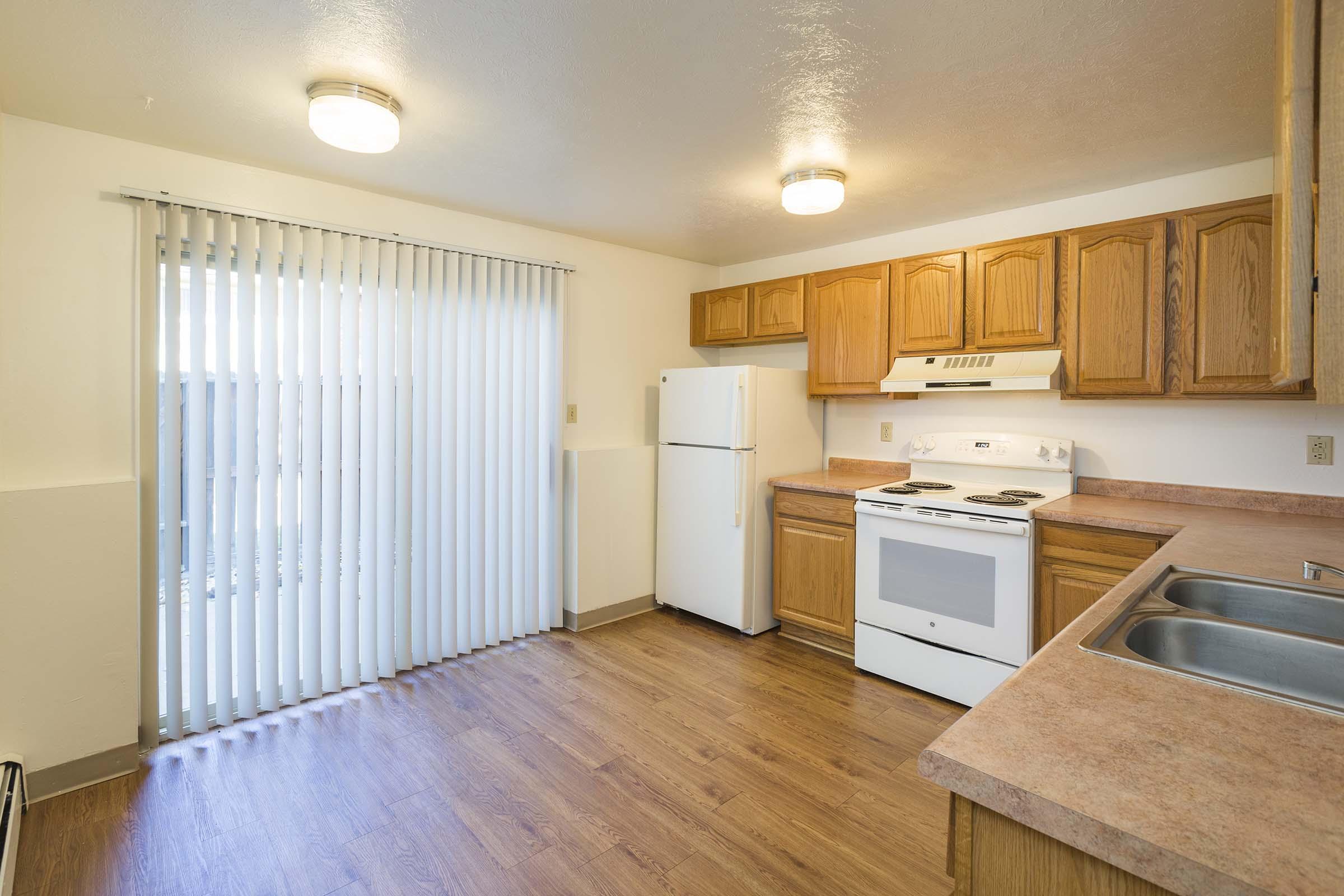 A bright kitchen featuring wooden cabinetry, a white refrigerator, and a white stove. Natural light streams in through sliding glass doors with vertical blinds, and the floor is covered in light wood laminate. A dual sink is visible on the counter, complementing the warm tones of the space.