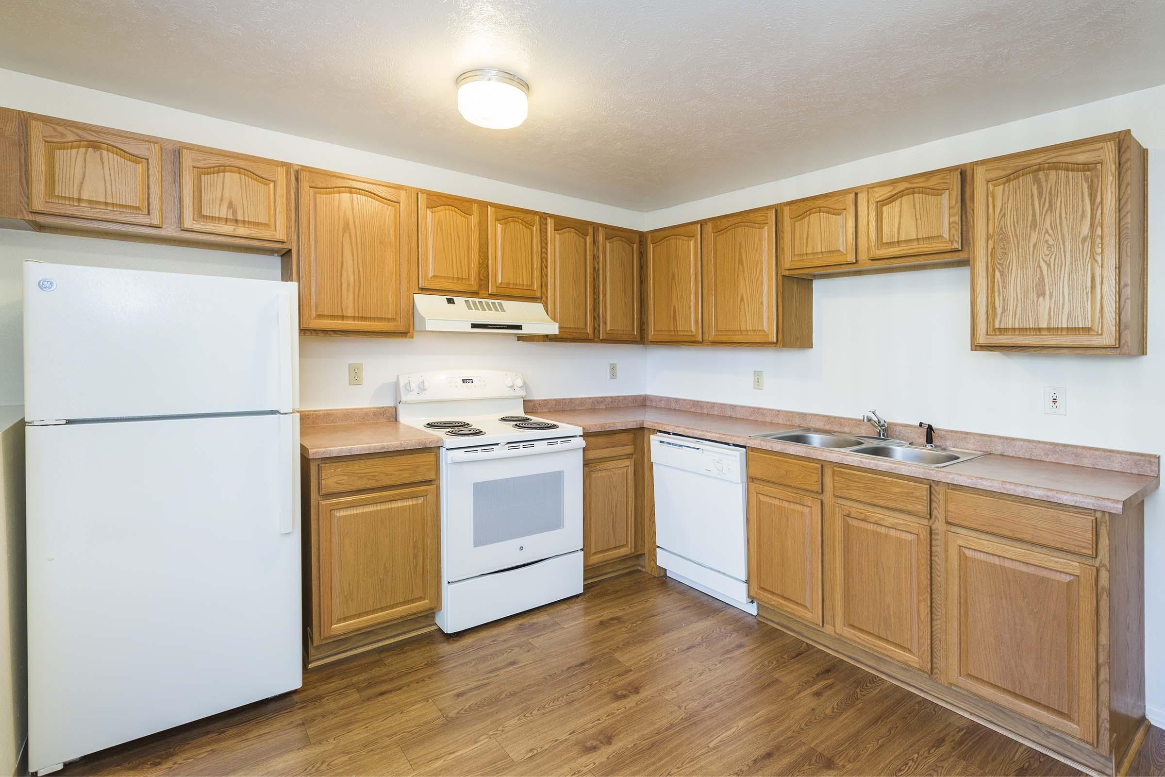 A modern kitchen featuring light wood cabinets, a white refrigerator, a white oven and stove, a double sink, and a dishwasher. The countertops are beige, and the floor is wooden laminate. Bright lighting illuminates the space, creating a warm and inviting atmosphere.