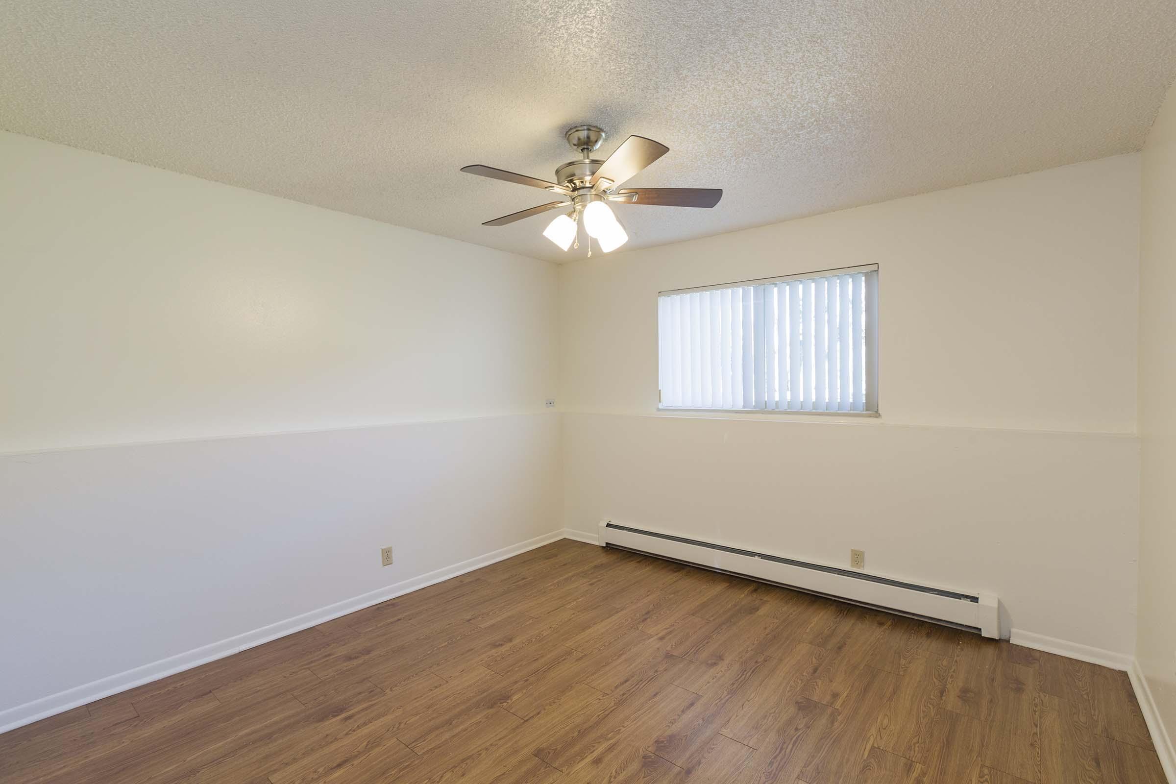 Empty room with light-colored walls and a ceiling fan. A window with vertical blinds allows natural light to enter. The floor is wooden, creating a warm atmosphere. There is a baseboard heater along one wall, and the space appears clean and ready for furnishing.