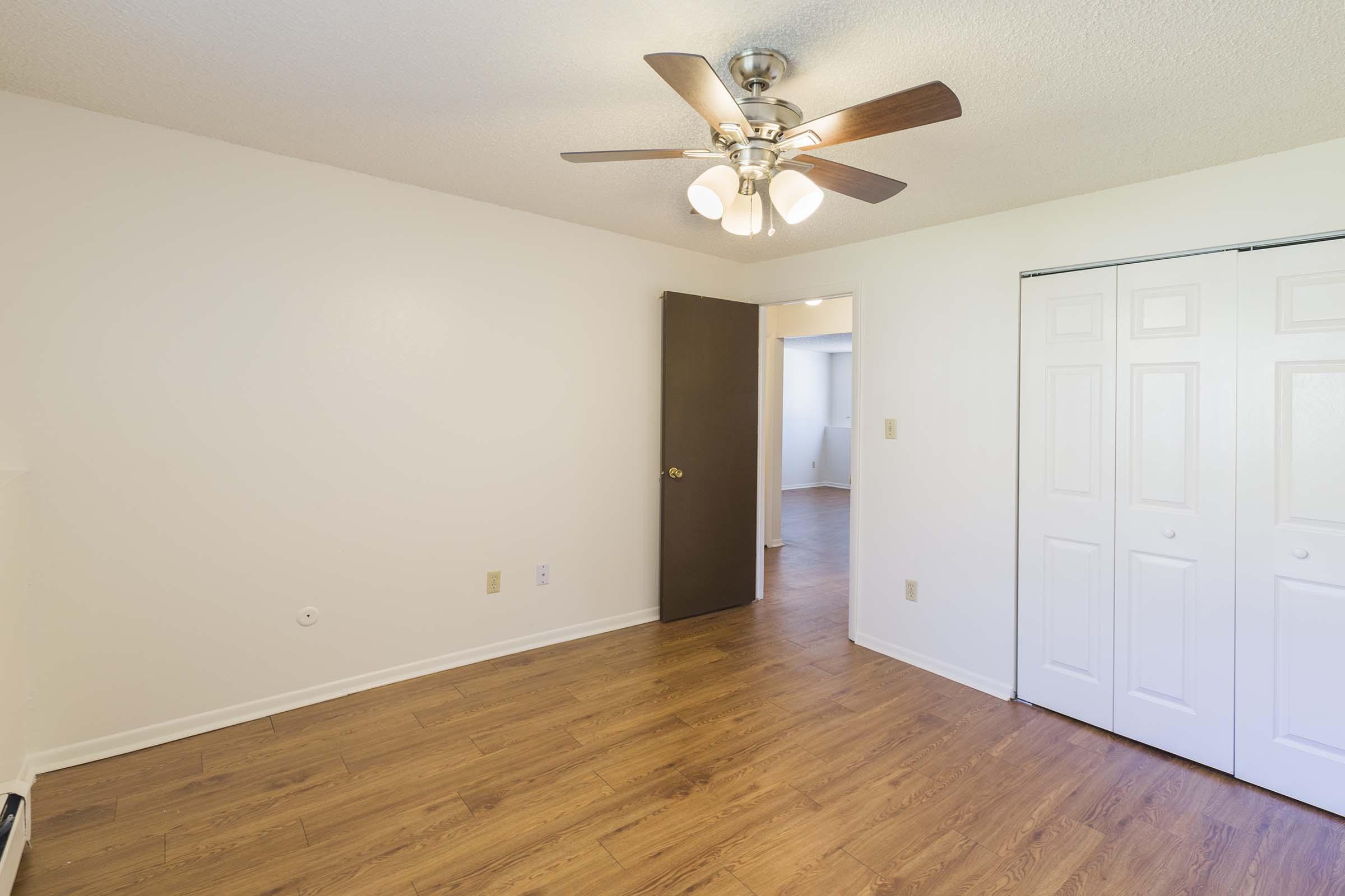 A well-lit room with hardwood-like floors, featuring a ceiling fan with four light bulbs. A closed brown door leads to another space, and there are double white closet doors. The walls are painted in a light color, creating a clean and open atmosphere.
