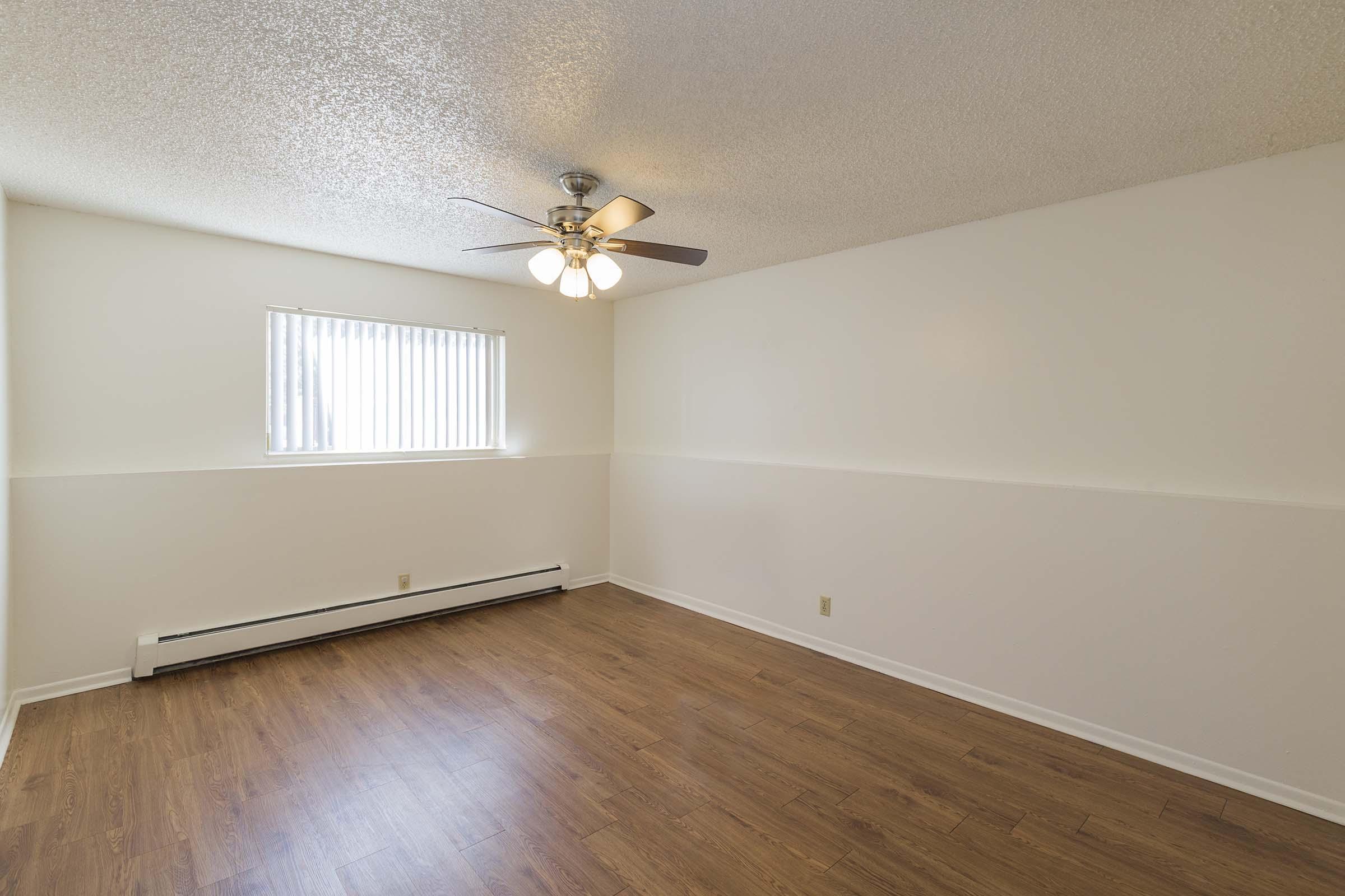 Empty room with light-colored walls and a textured ceiling. A ceiling fan with three lights is visible. A window with vertical blinds allows natural light to enter, and the floor is covered with laminate in a warm tone. Baseboard heating is present along one wall.