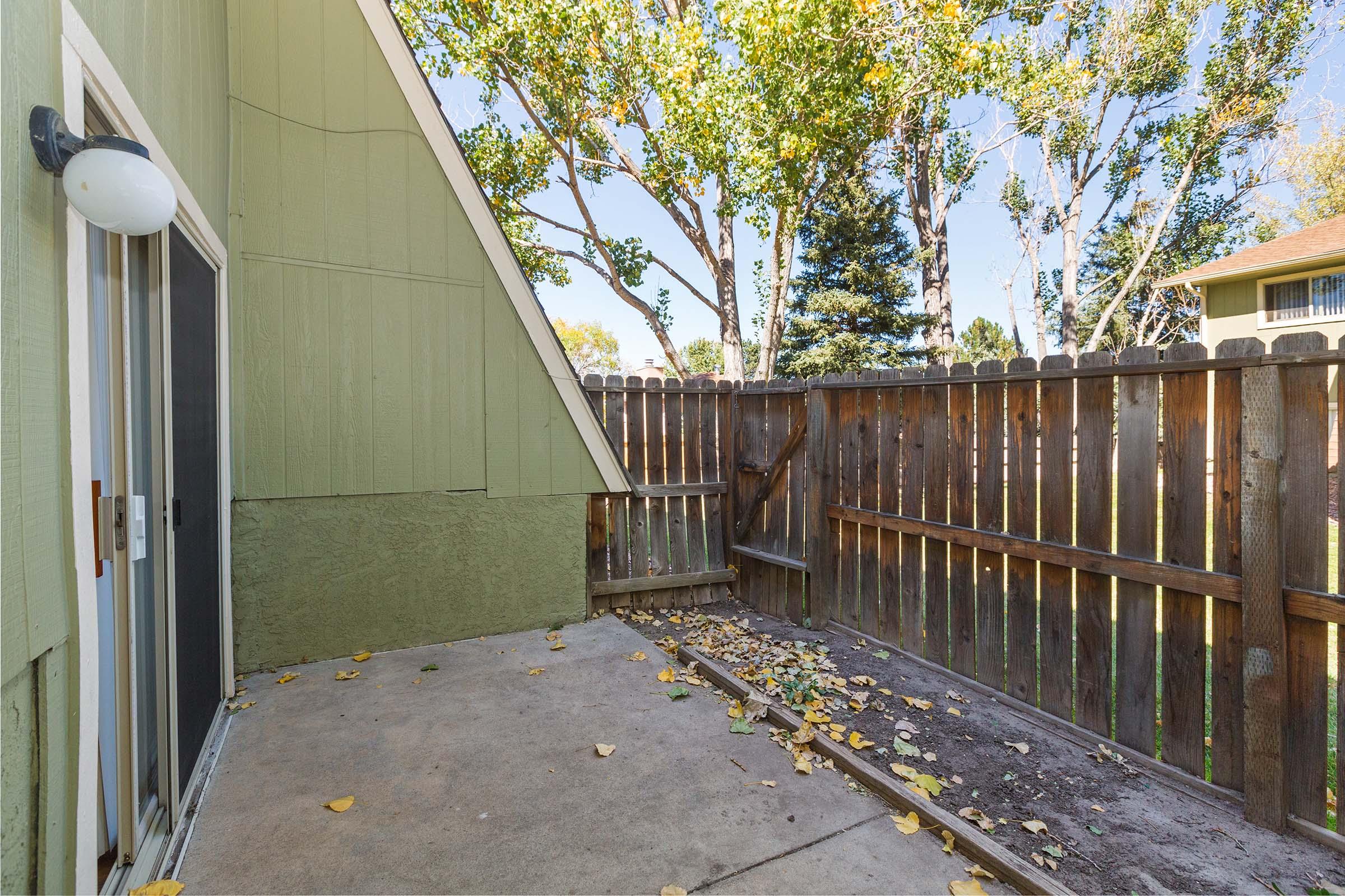 A small outdoor patio area with a concrete surface, featuring a wooden fence and autumn leaves scattered on the ground. Tall trees are visible in the background, and there is a light fixture mounted on the wall of a green building. The space appears secluded and quiet.