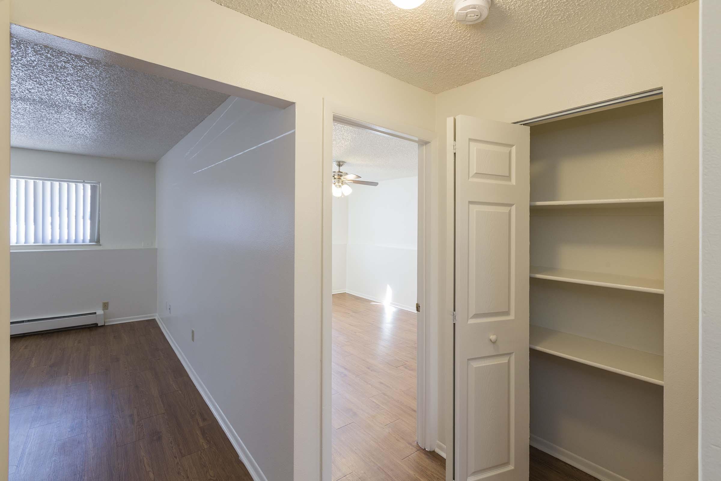 Interior view of a light-colored apartment hallway with a doorway leading to a closet on the right. The room features a window with blinds and a ceiling fan. The flooring is wood-like, and there is a bright, open space visible in the background.