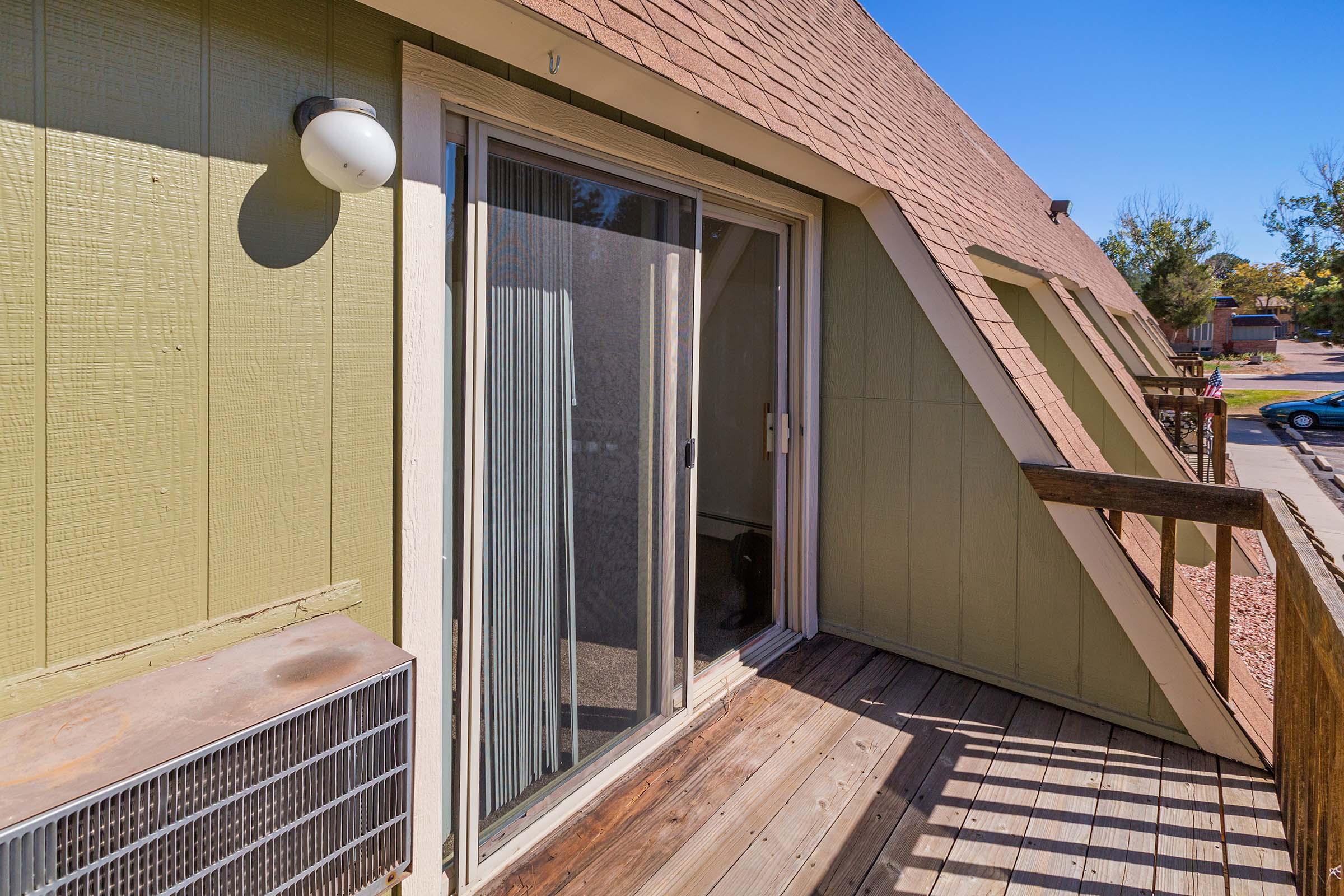 Sliding glass door leading to a small balcony area with wooden railing, situated in a light green building with a brown, sloped roof. An air conditioning unit is mounted on the exterior wall near the door, and the view outside shows a pathway and trees in the background.
